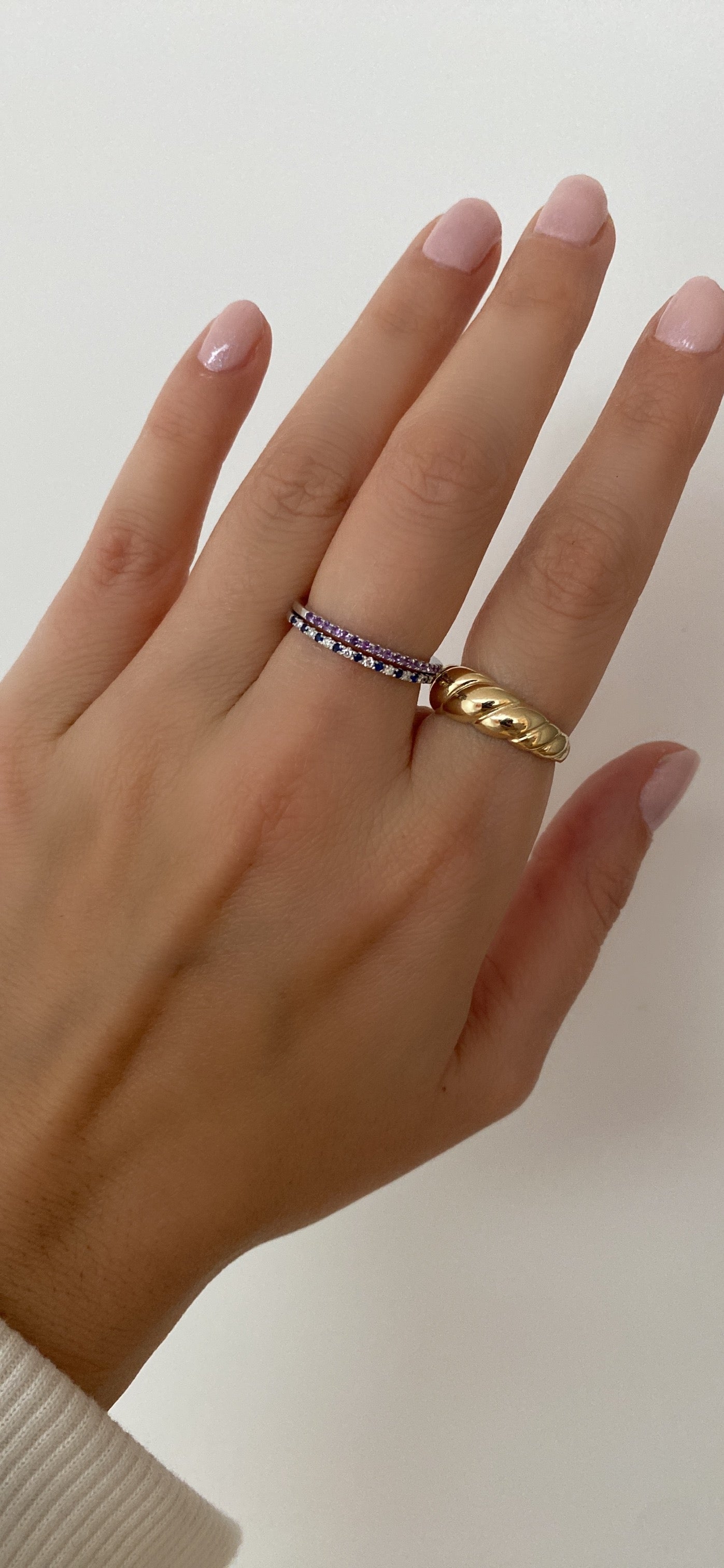 A person's hand wearing an alternating diamond and birthstone ring styled with other diamond, coloured stone, and gold ring against a neutral background.
