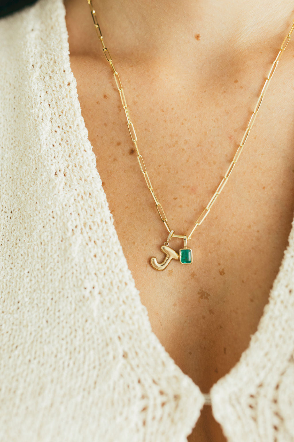 close up of person wearing a paperclip necklace with a diamond puffy letter and emerald pendant attached to it