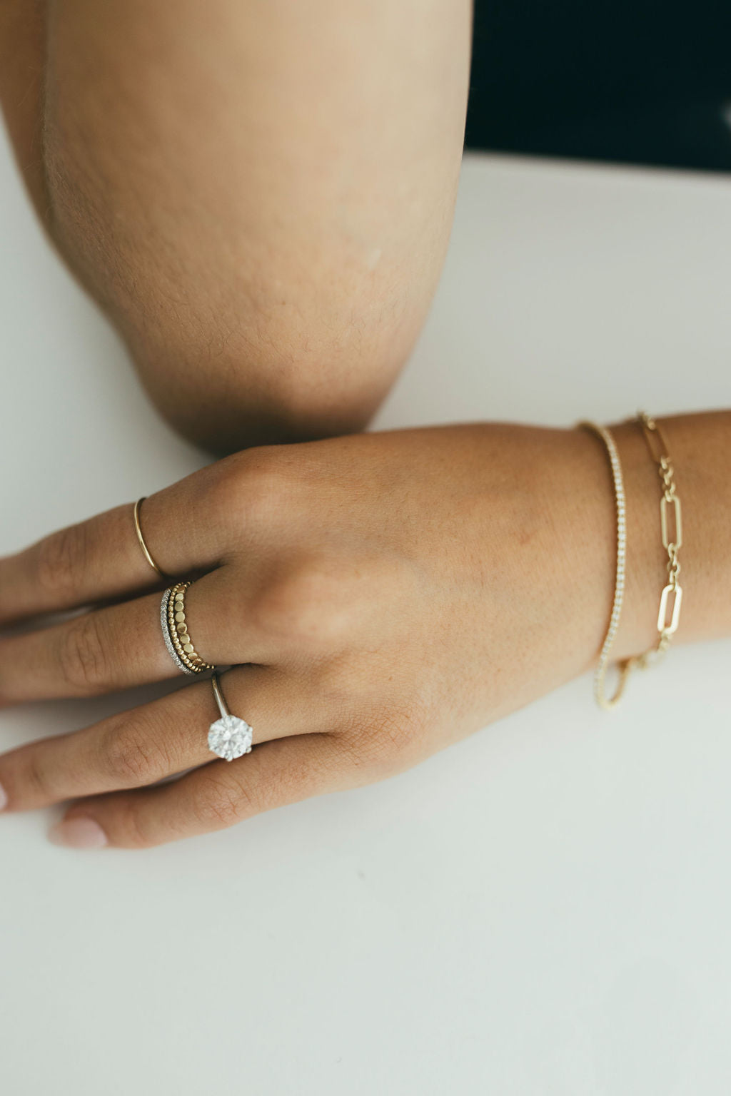 A hand wearing a diamond ring with other gold rings and dainty bracelets on a light background.