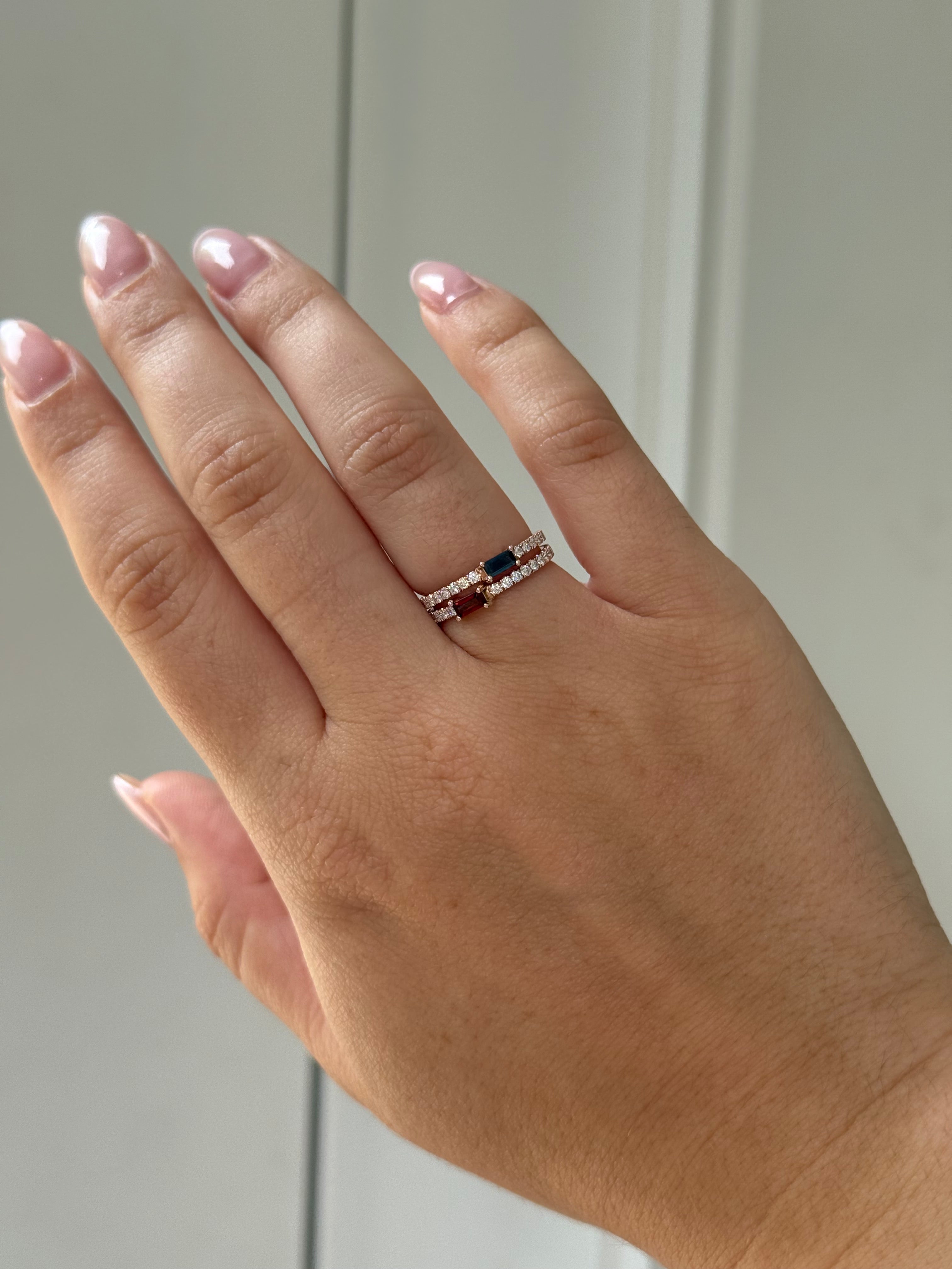 Hand wearing two baguette birthstone and diamond rings on a neutral background.