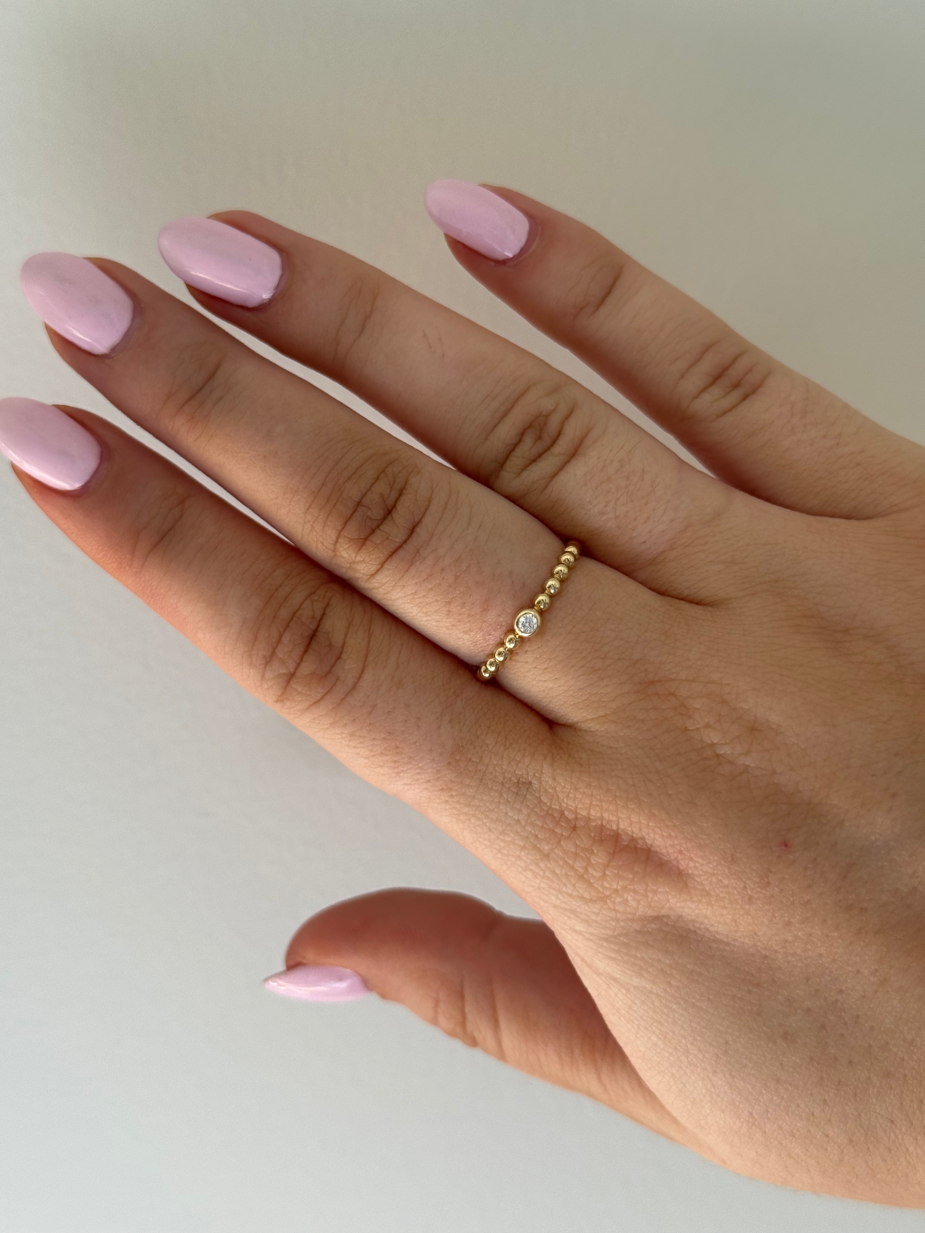 Hand wearing a gold bead ring with a single diamond on a neutral background.