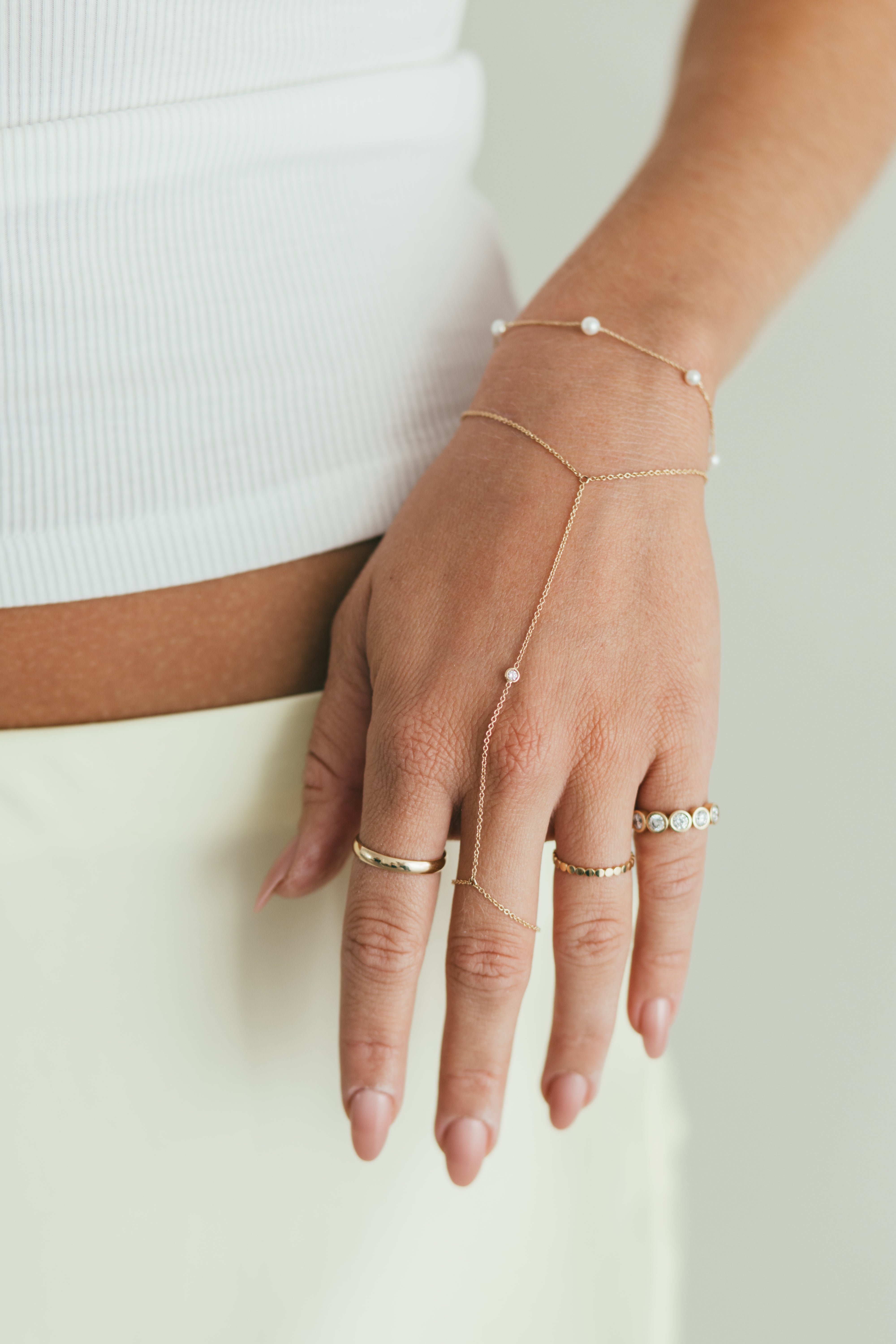A person's hand wearing a gold diamond hand chain and multiple gold rings, styled with a white top and pastel skirt.