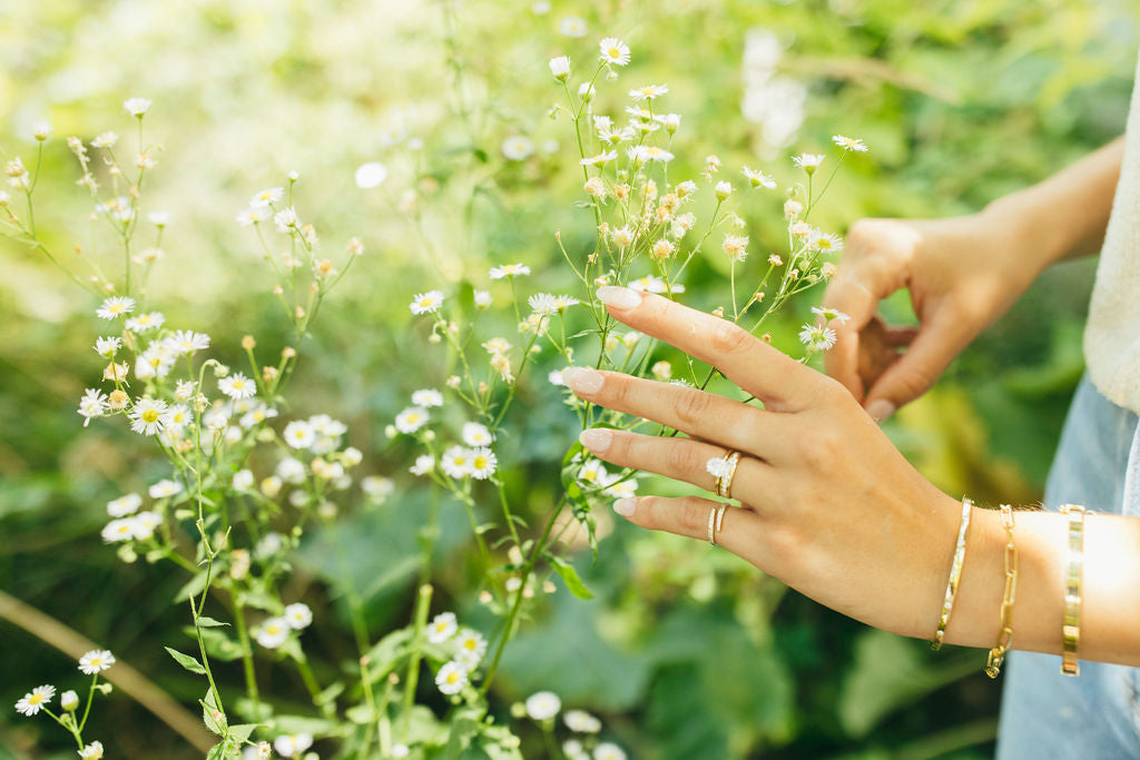 A hand styled with a mix of gold and diamond rings touching delicate flowers in a natural setting.