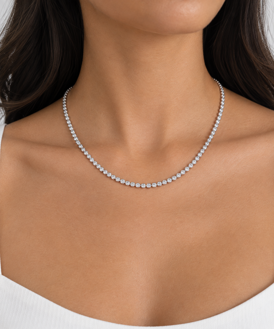 A close-up of a model wearing a bezel-set white gold tennis necklace against a clean white background.