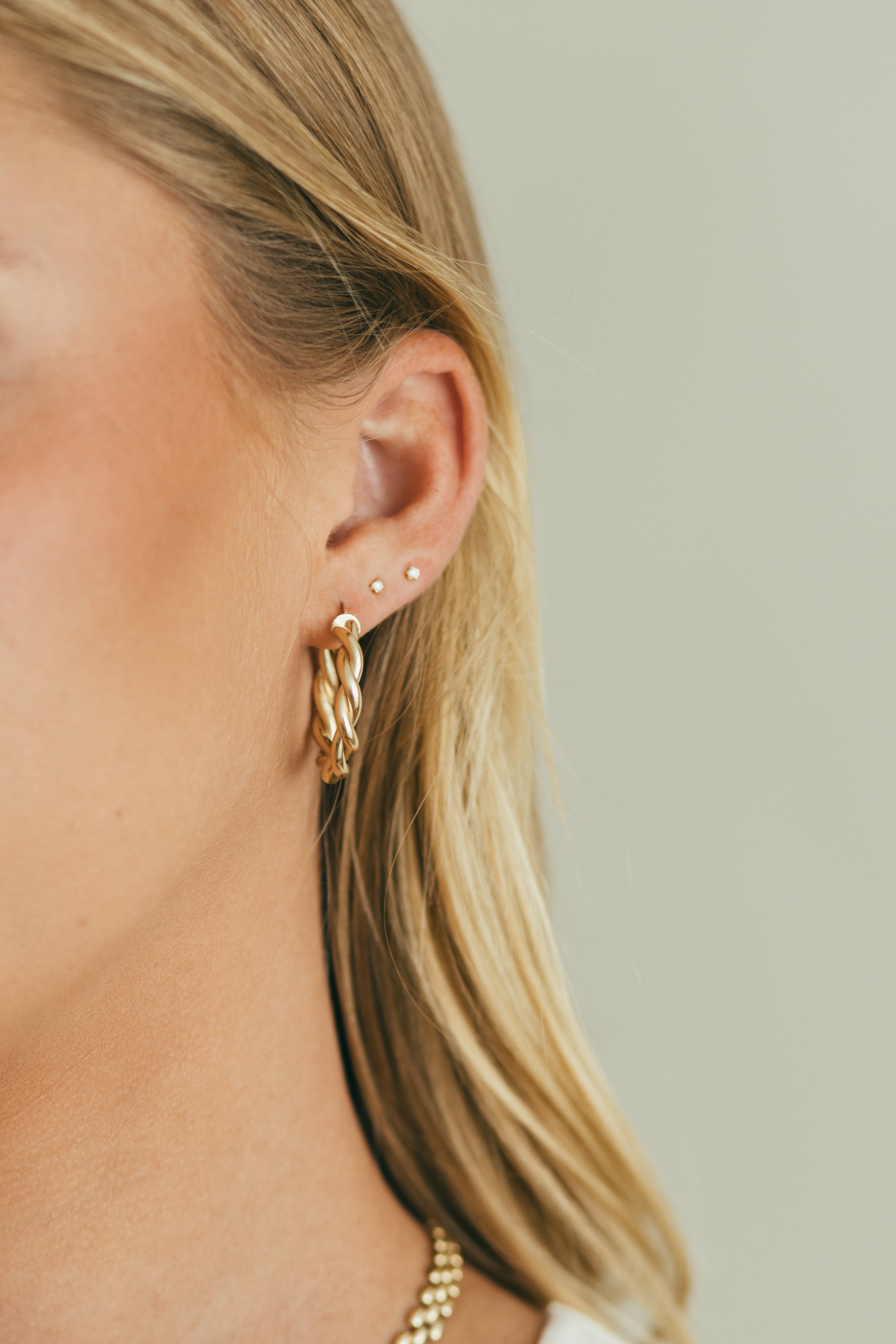 Close-up of a person wearing gold twisted hoop earrings and diamond studs with a neutral background
