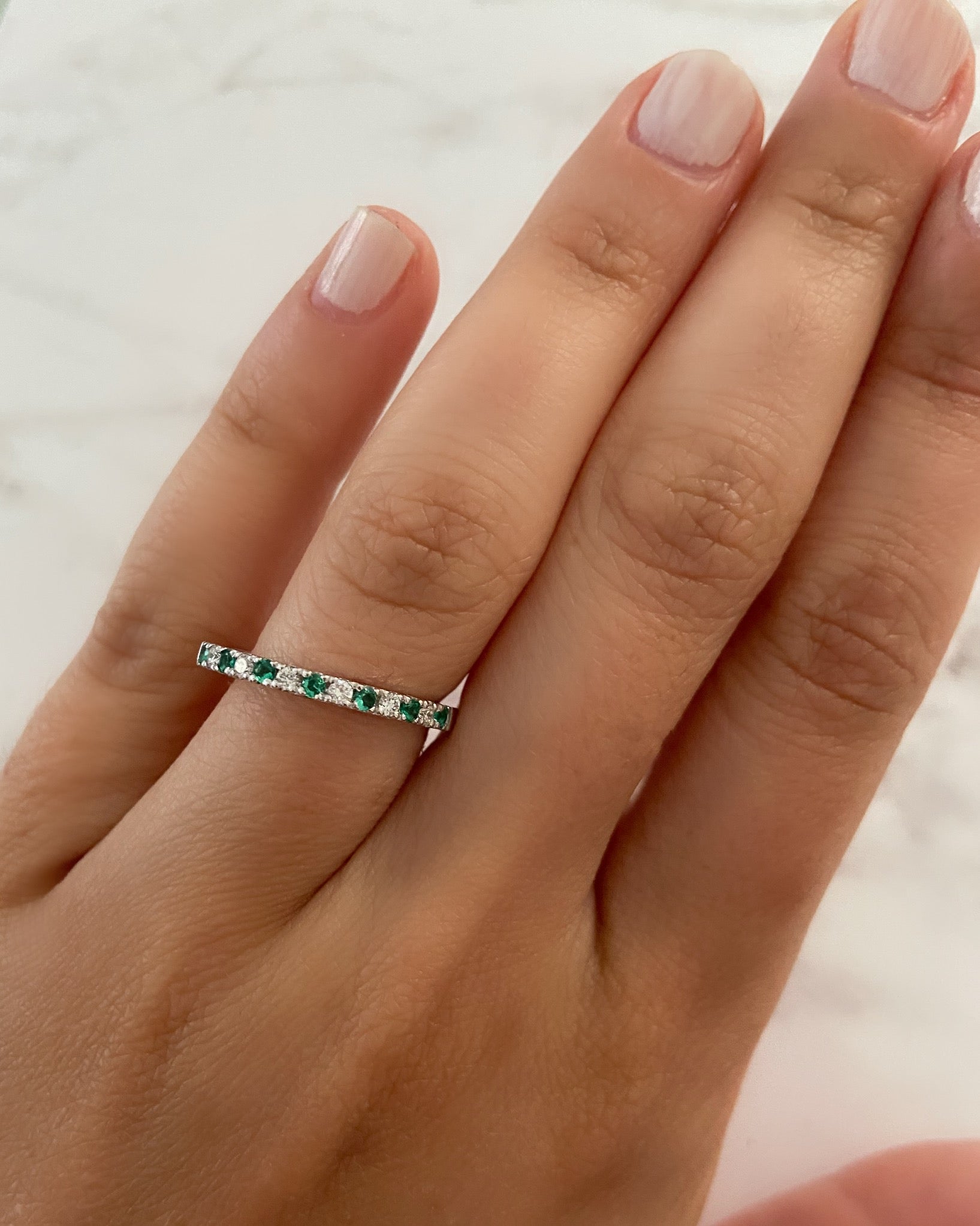 A person's hand wearing an alternating diamond and birthstone ring against a neutral background.