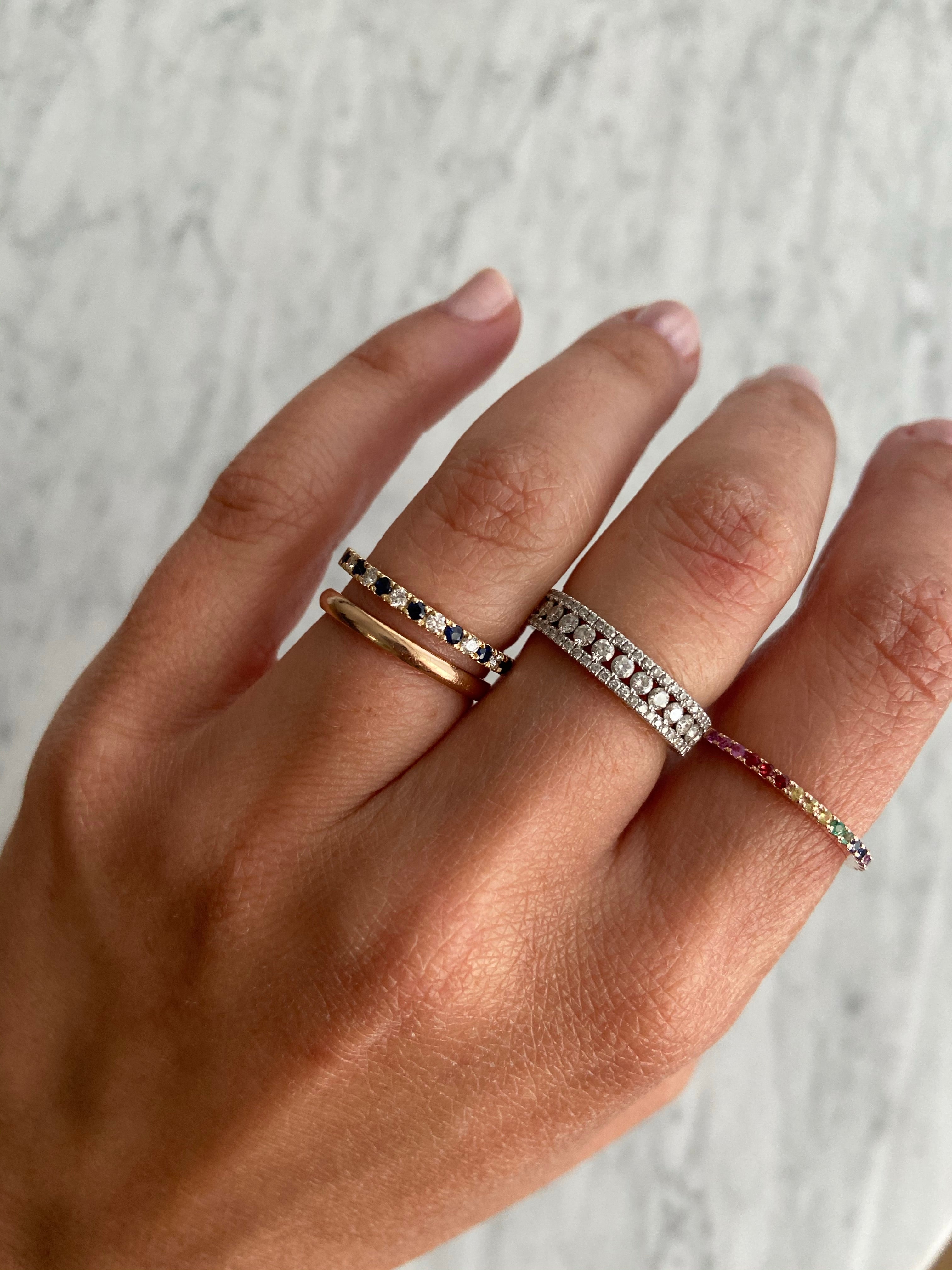 A person's hand wearing an alternating diamond and birthstone ring styled with other diamond, coloured stone, and gold rings against a neutral background.