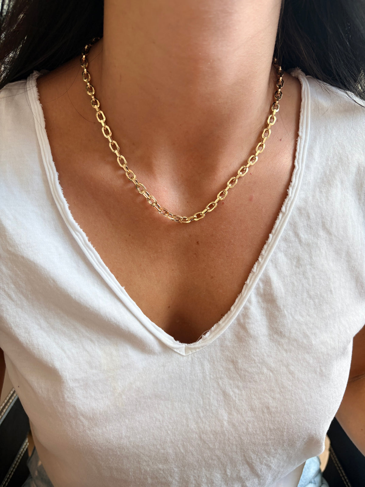 A close-up of a person wearing a gold chain necklace with interlocking oval links, styled with a white V-neck t-shirt.