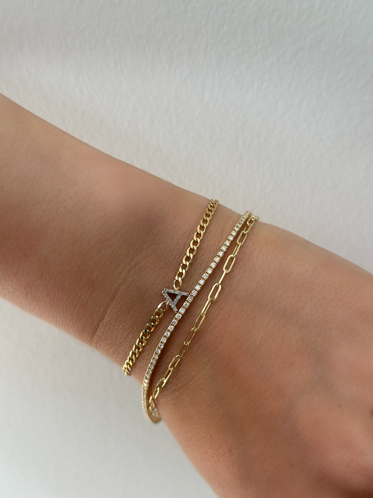 Curb chain bracelet with a diamond letter attached, stacked with a gold paperclip bracelet and diamond tennis bracelet.