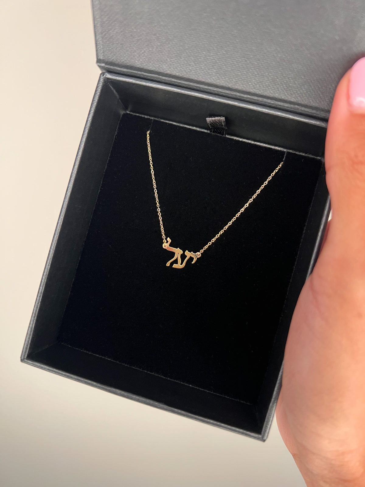 a yellow gold Hebrew name necklace in block font styled, placed in a black cushion box and held against a neutral background.