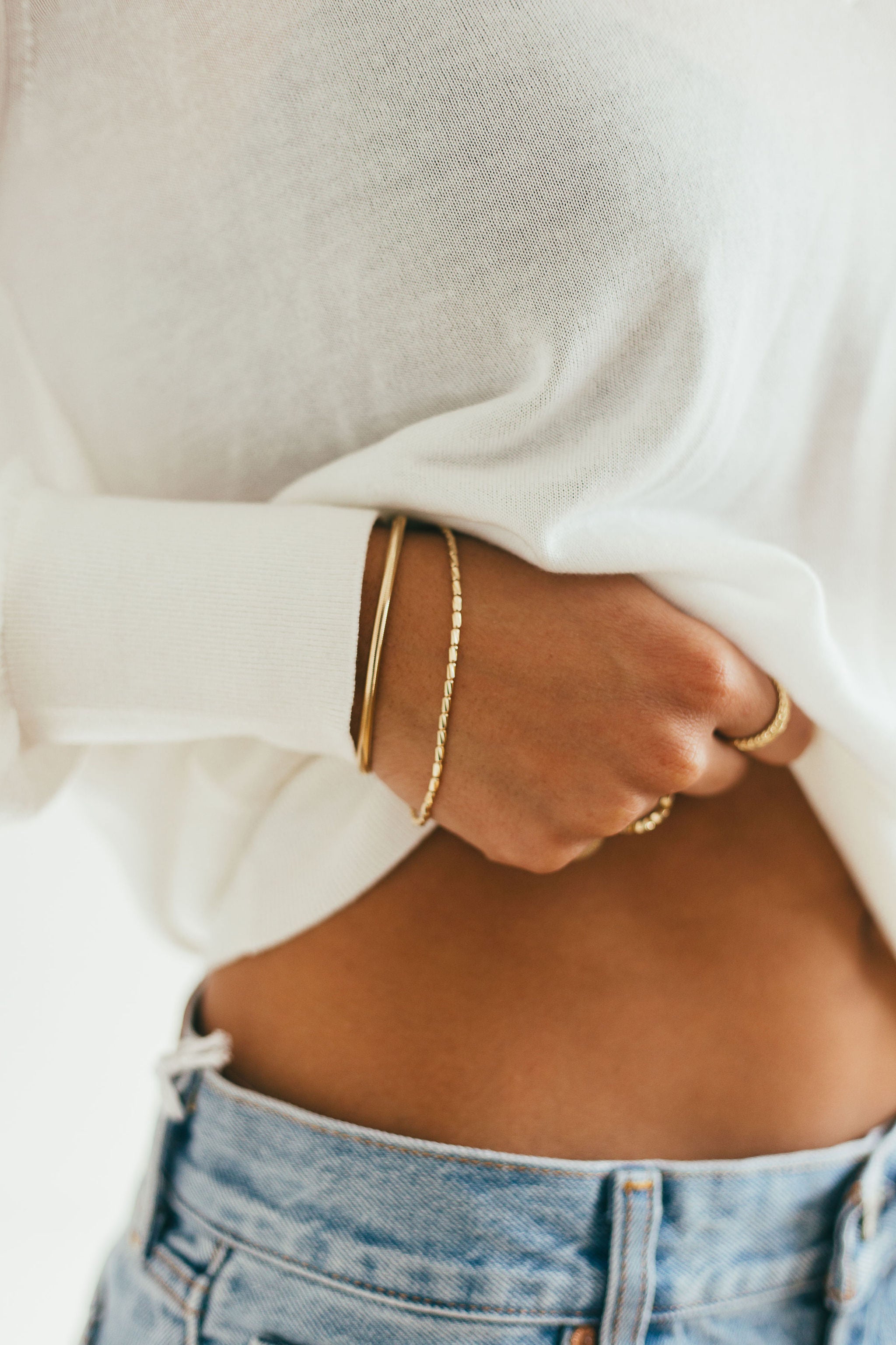 Person wearing a white sweater and blue jeans with gold jewelry on a neutral background