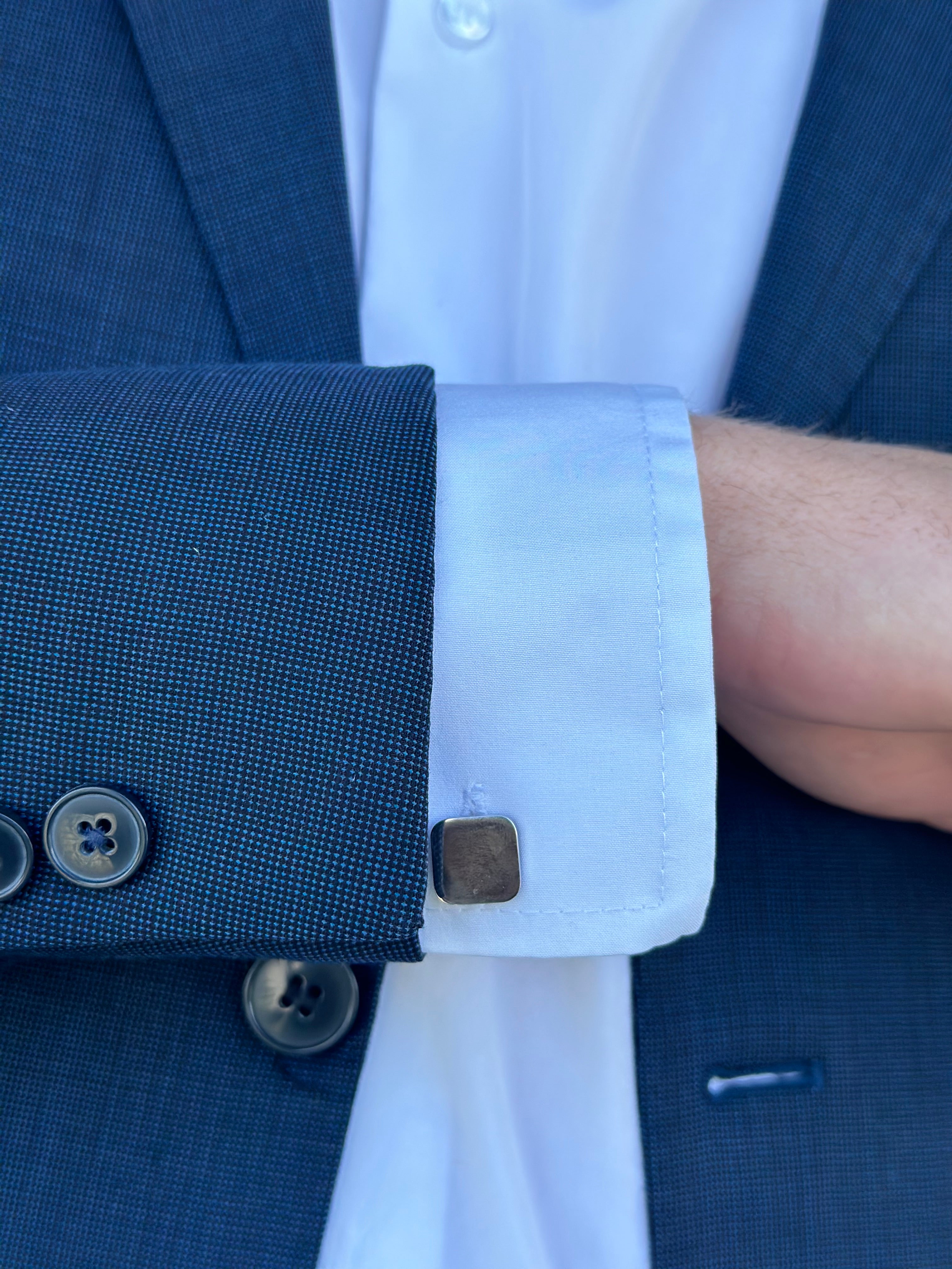 A square stainless steel cufflink worn on the cuff of a man's dress shirt with a blue suit jacket.