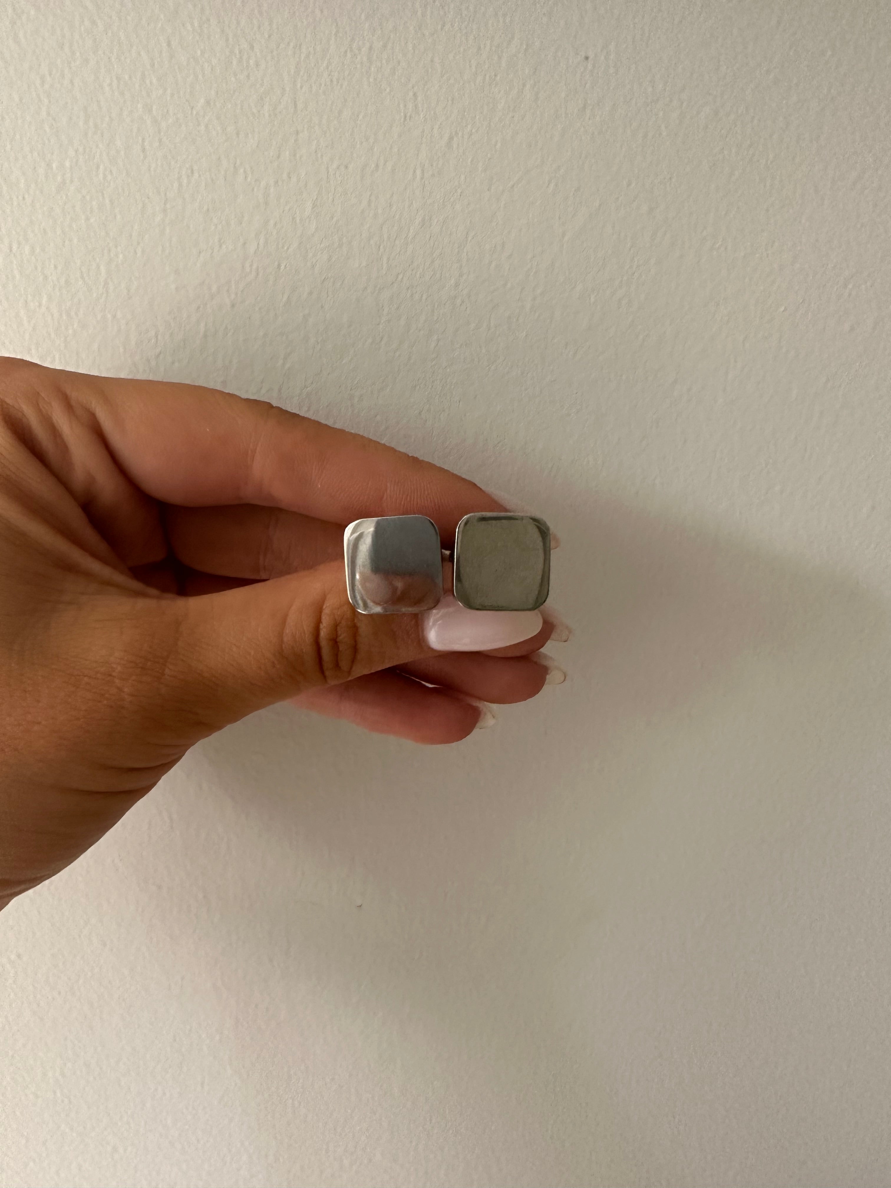 A hand holding a single square-shaped stainless steel cufflink against a neutral background.