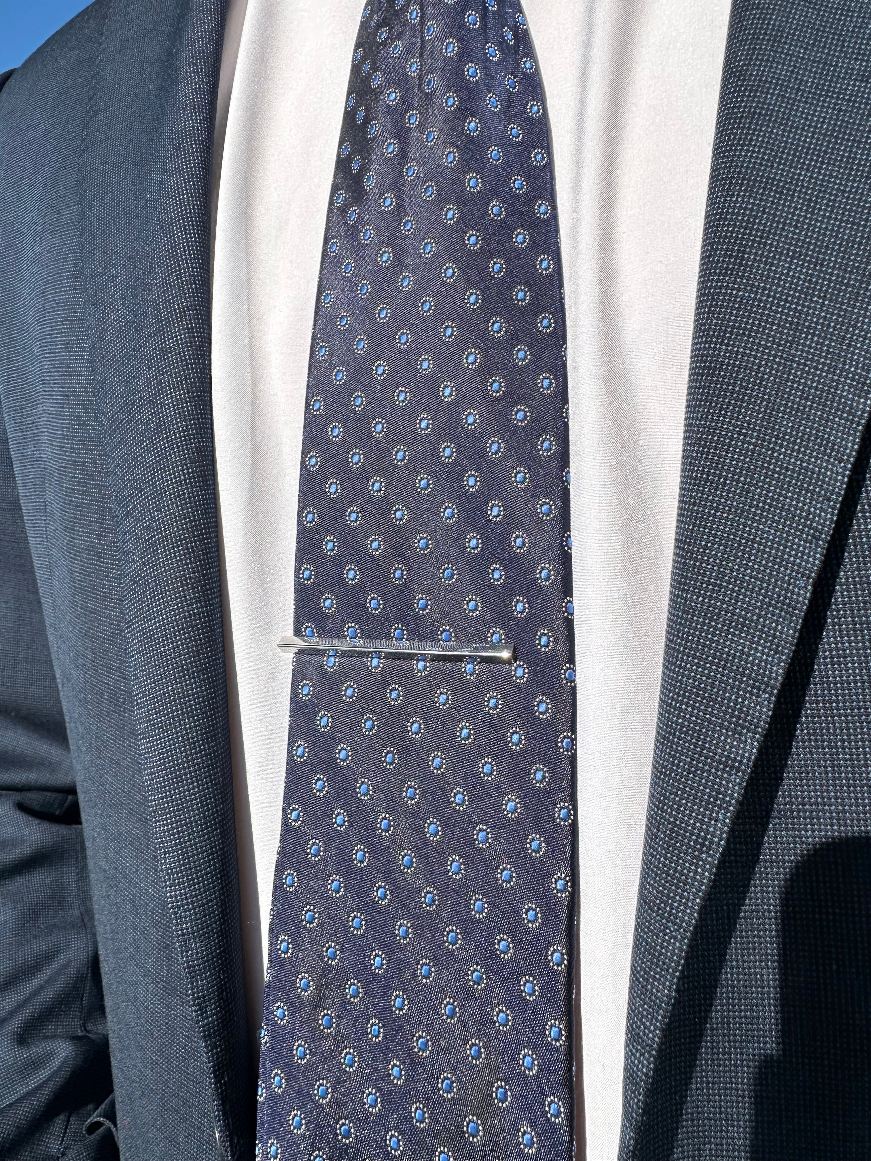 A sterling silver triangular tie bar worn on a men's dark blue patterned tie with a white dress shirt and dark blue suit jacket.