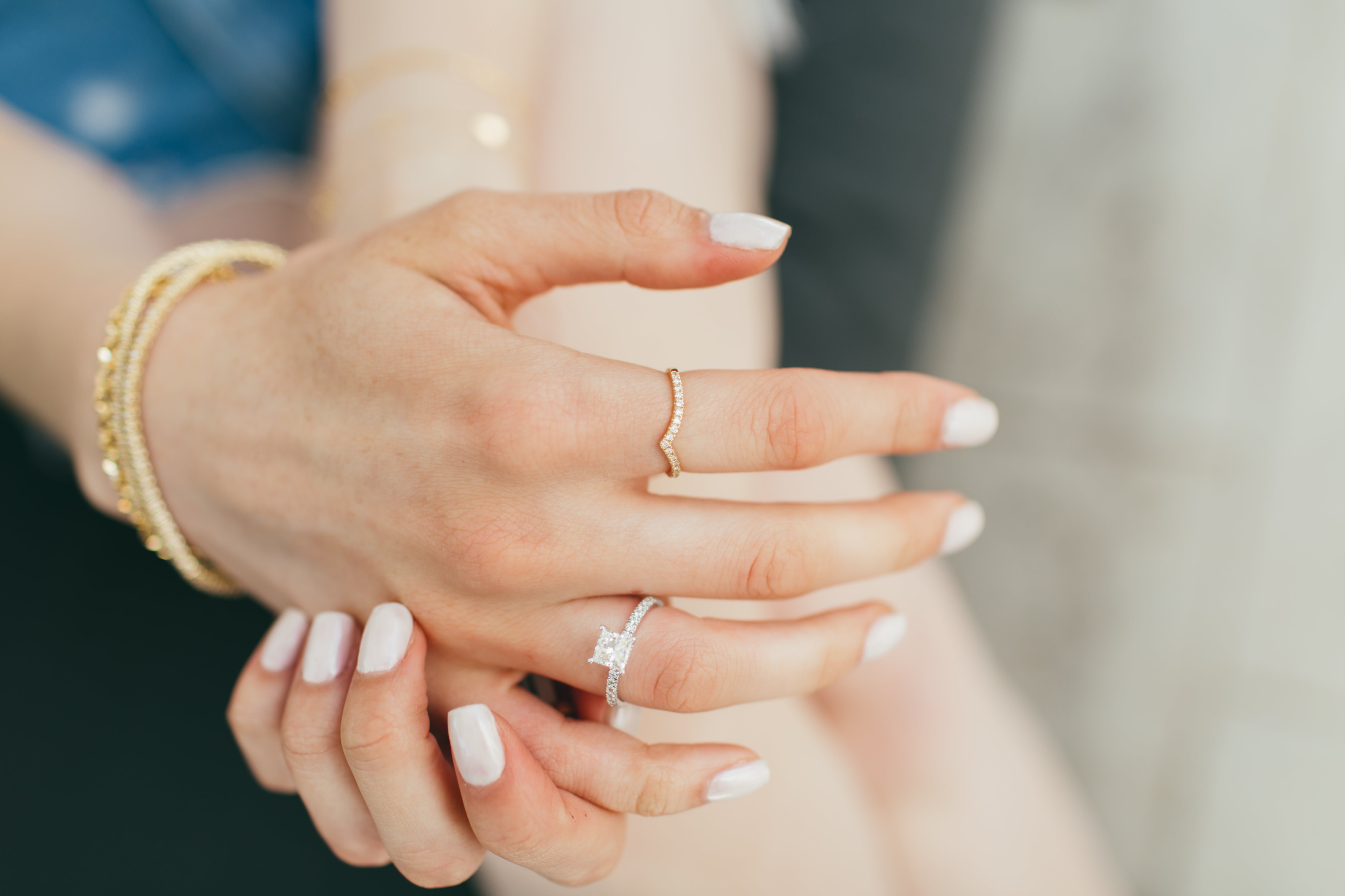 A close-up of a woman's hands styled with a diamond v ring nad other diamond ring and gold bracelets on a blurred background.