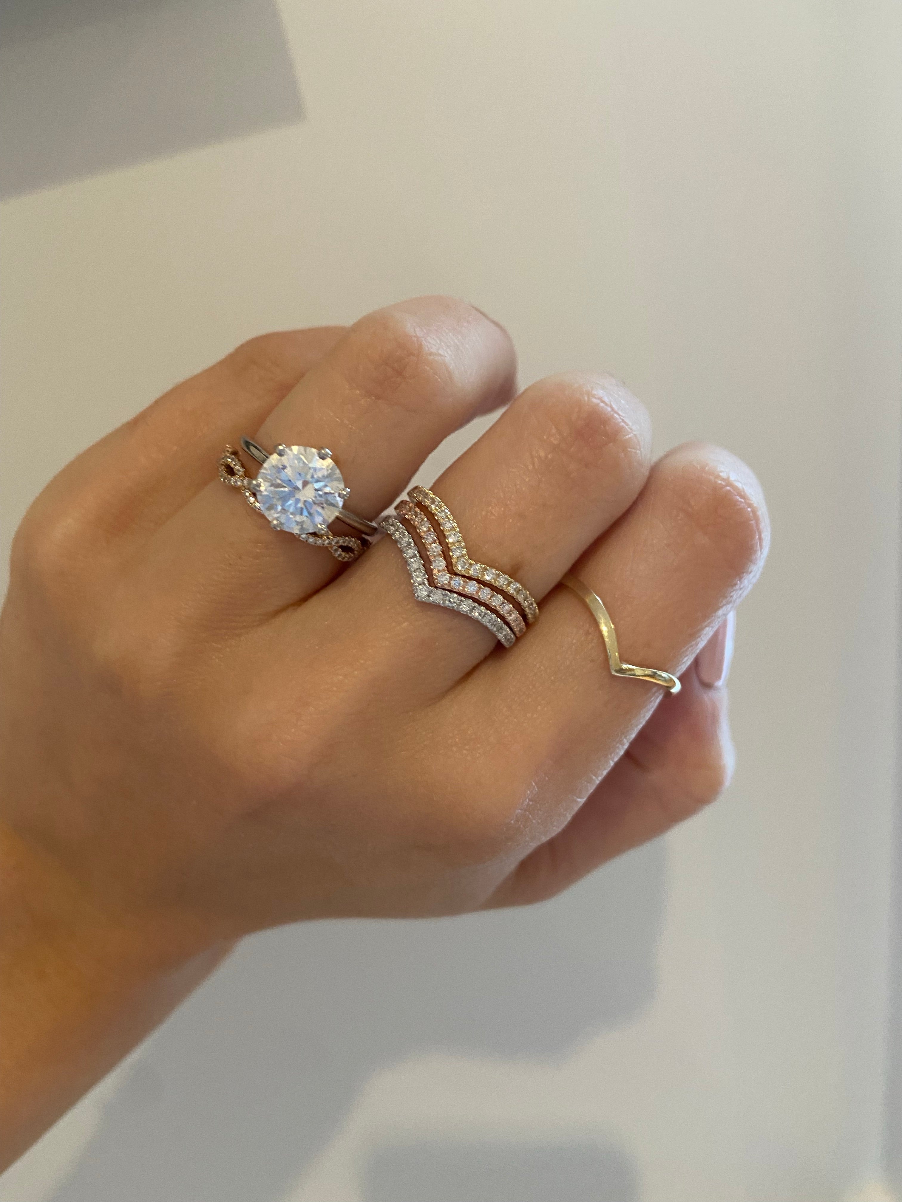 A close up of a hand wearing a stack of yellow gold, rose gold, and white gold diamond v rings on a neutral background.
