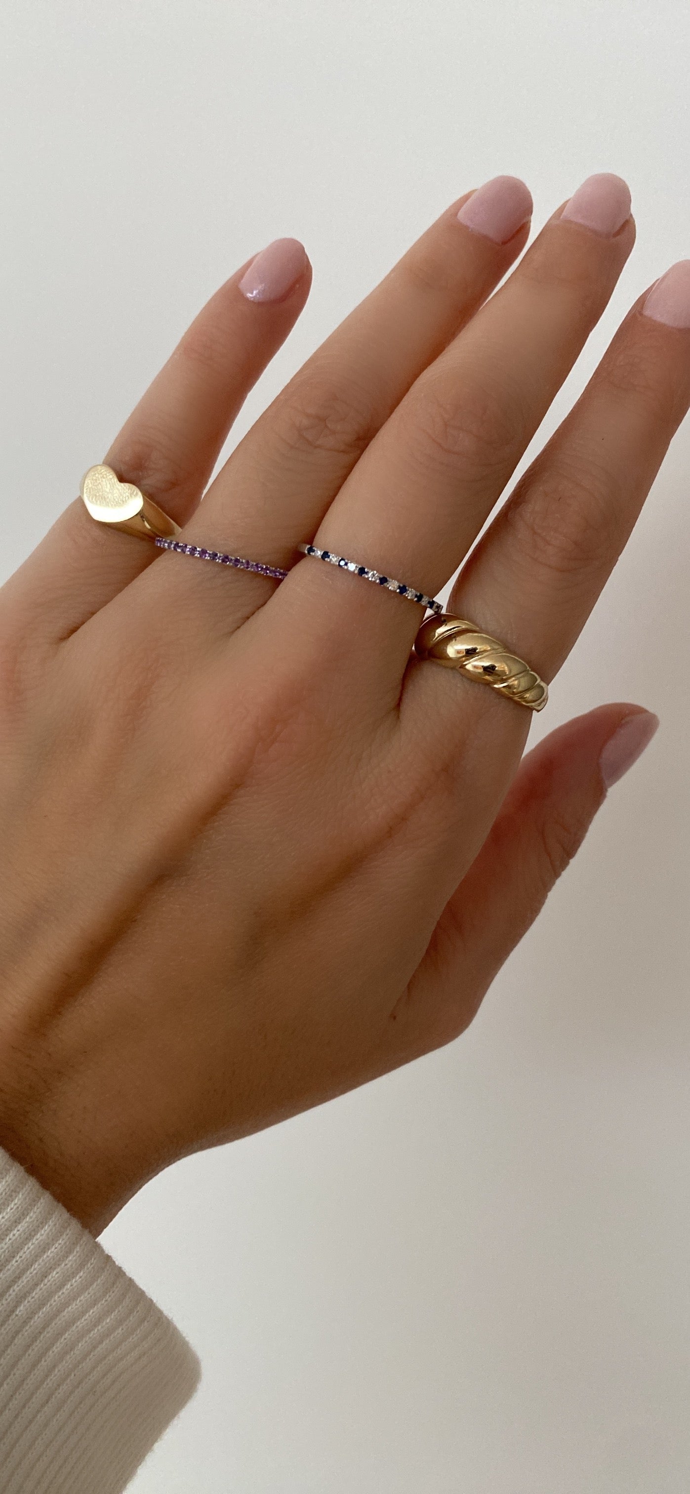 A person's hand wearing an alternating diamond and birthstone ring styled with other diamond, coloured stone, and gold rings against a neutral background.