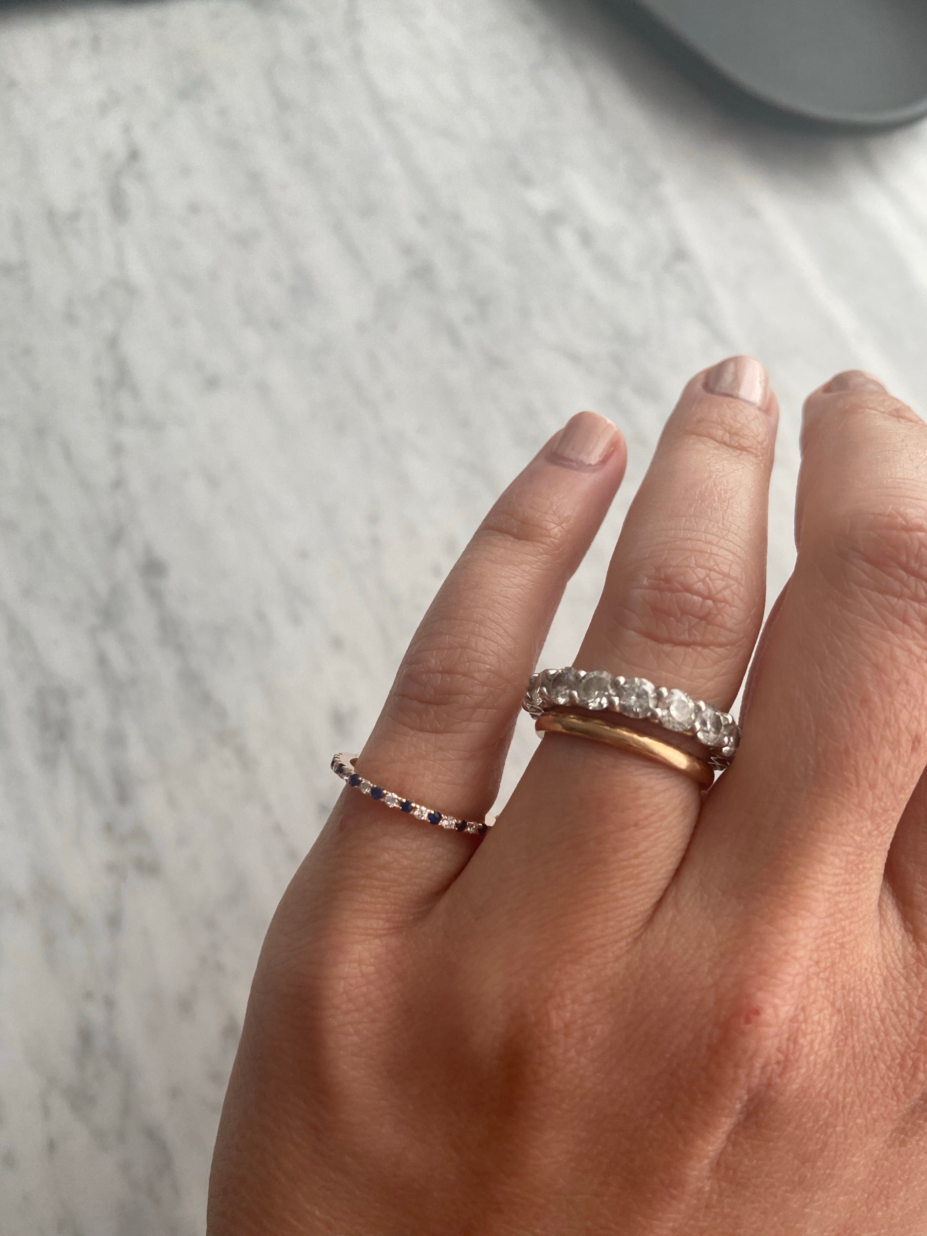 A person's hand wearing an alternating diamond and birthstone ring styled with a diamond and gold ring against a neutral background.