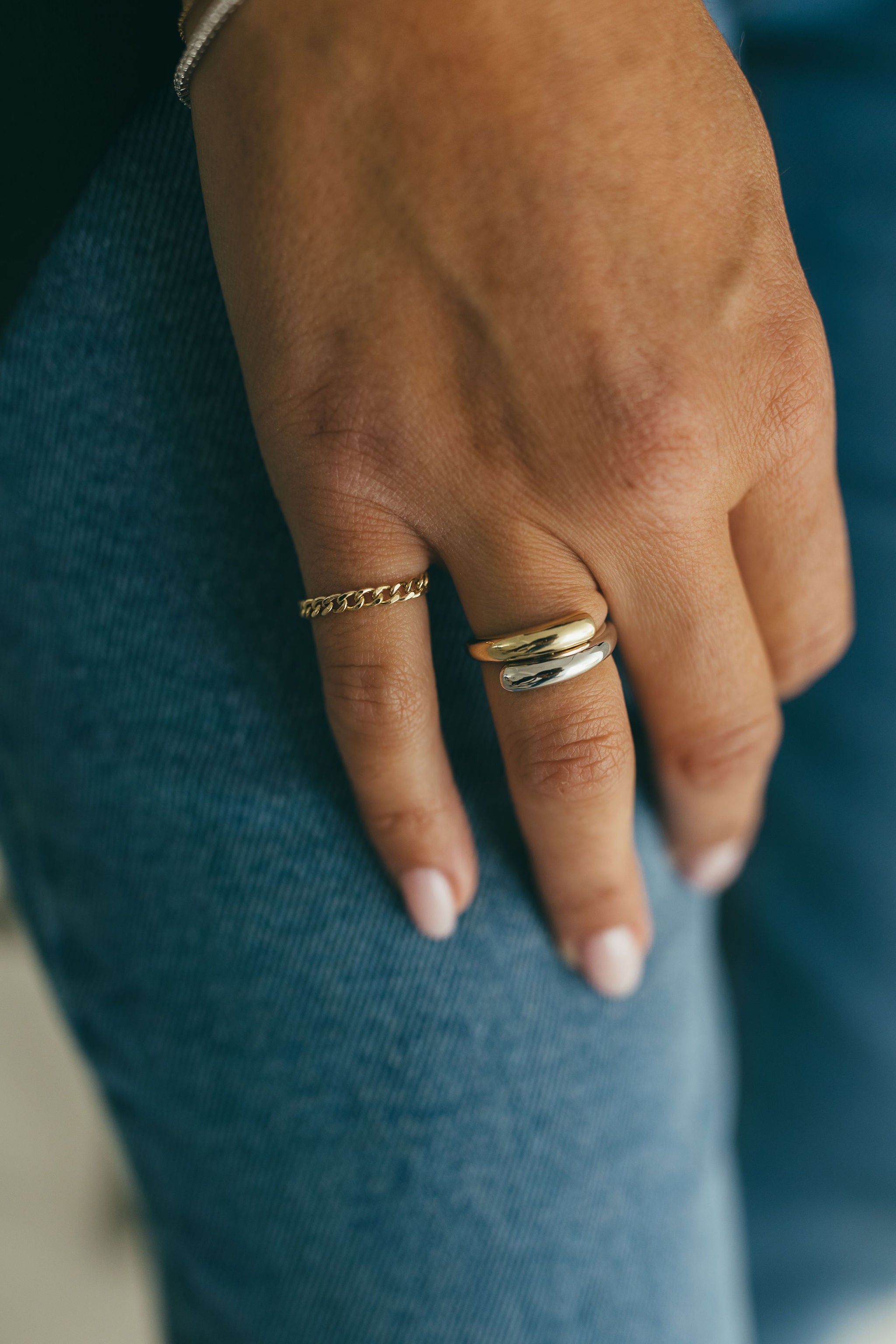 A chunky white and yellow gold wrap ring styled with a gold ring on a person's hand against denim jeans.