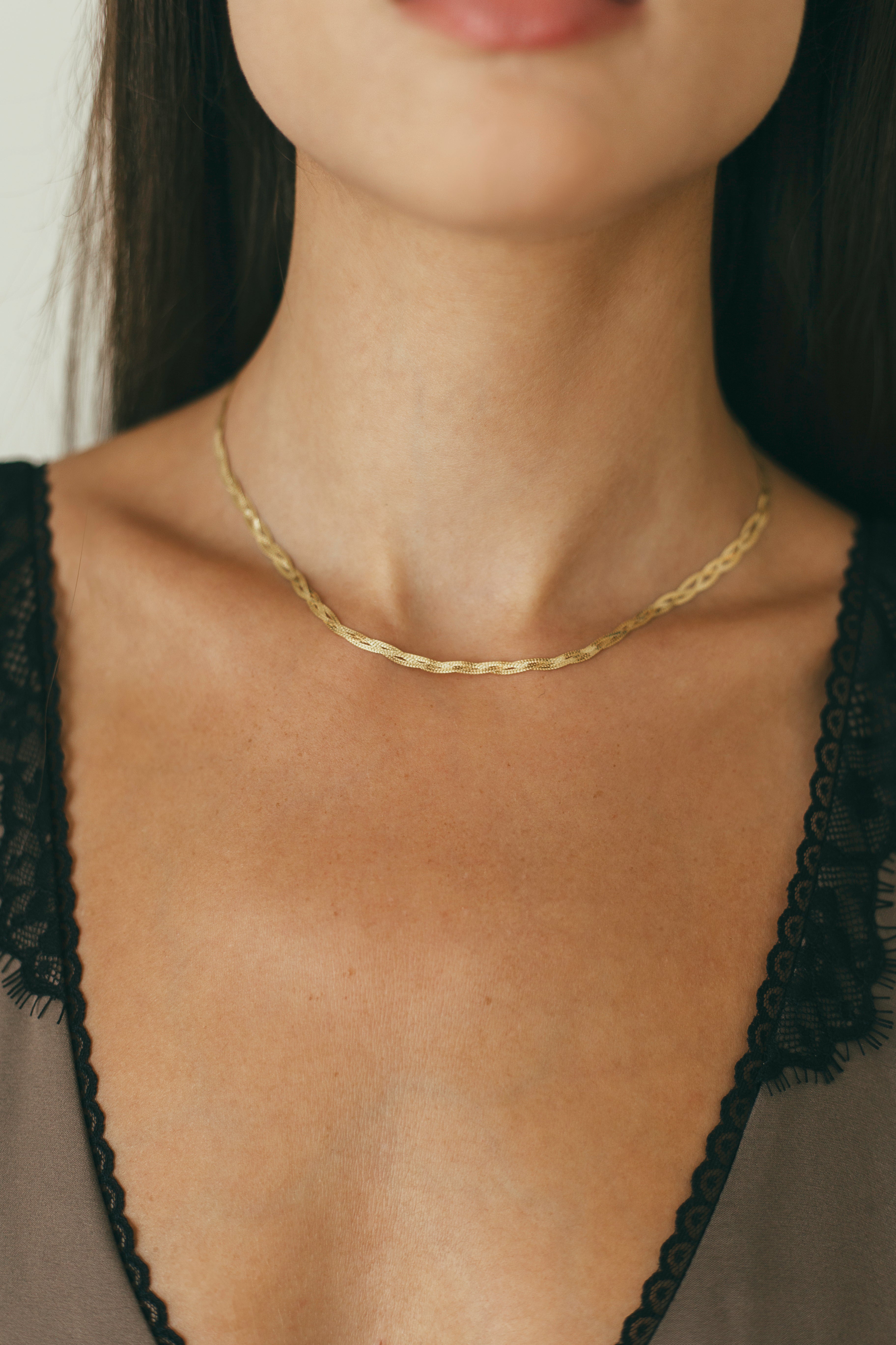 A close up of a gold braided herringbone necklace on a person wearing a black lace top.