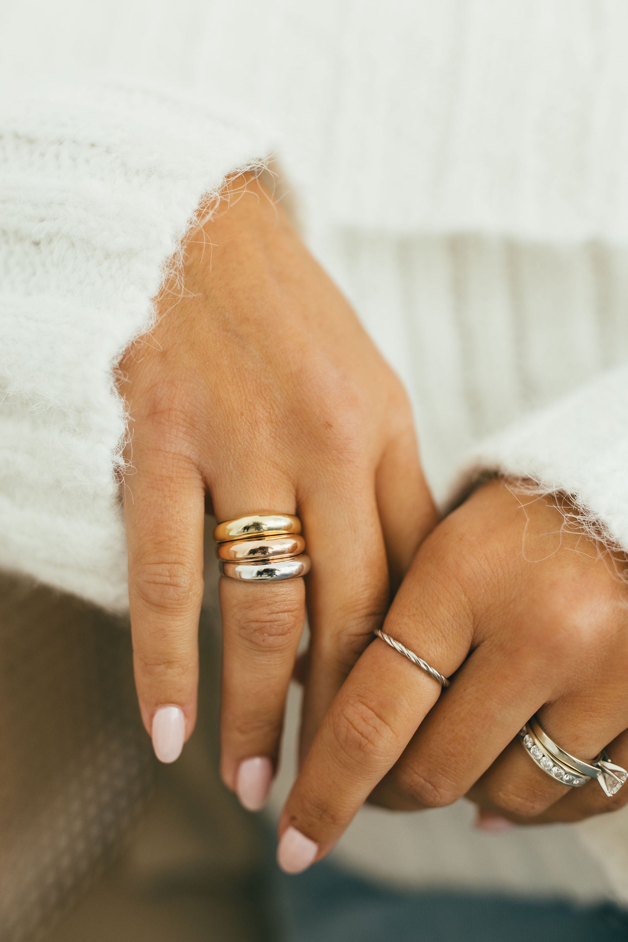 A close up of a woman's hands wearing a stack of three chunky dome rings in yellow gold, rose gold, and white gold, styled with a fuzzy white sweater.