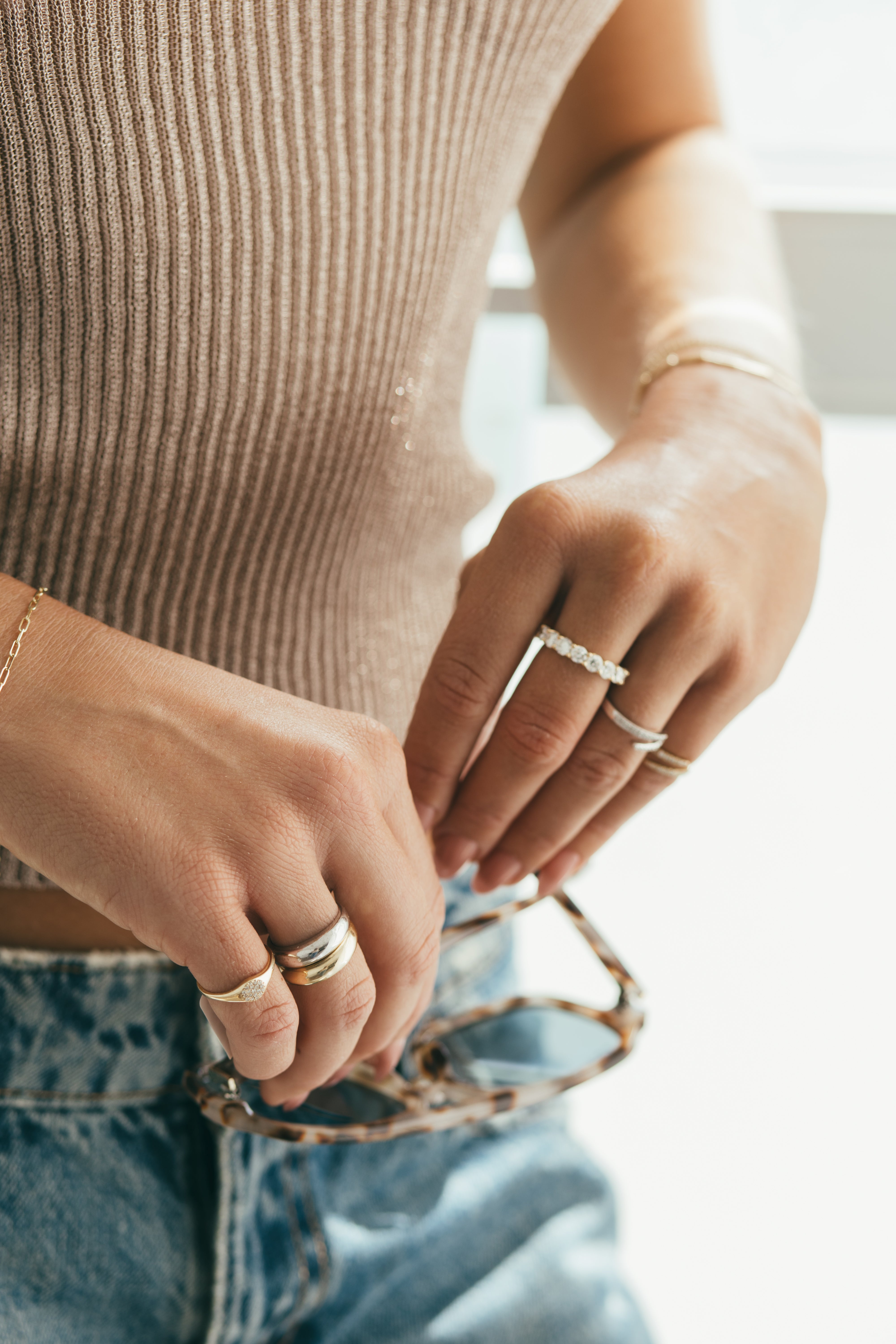 A close up of a woman's hands styled with a mix of gold and diamond rings while holding a pair of sunglasses.