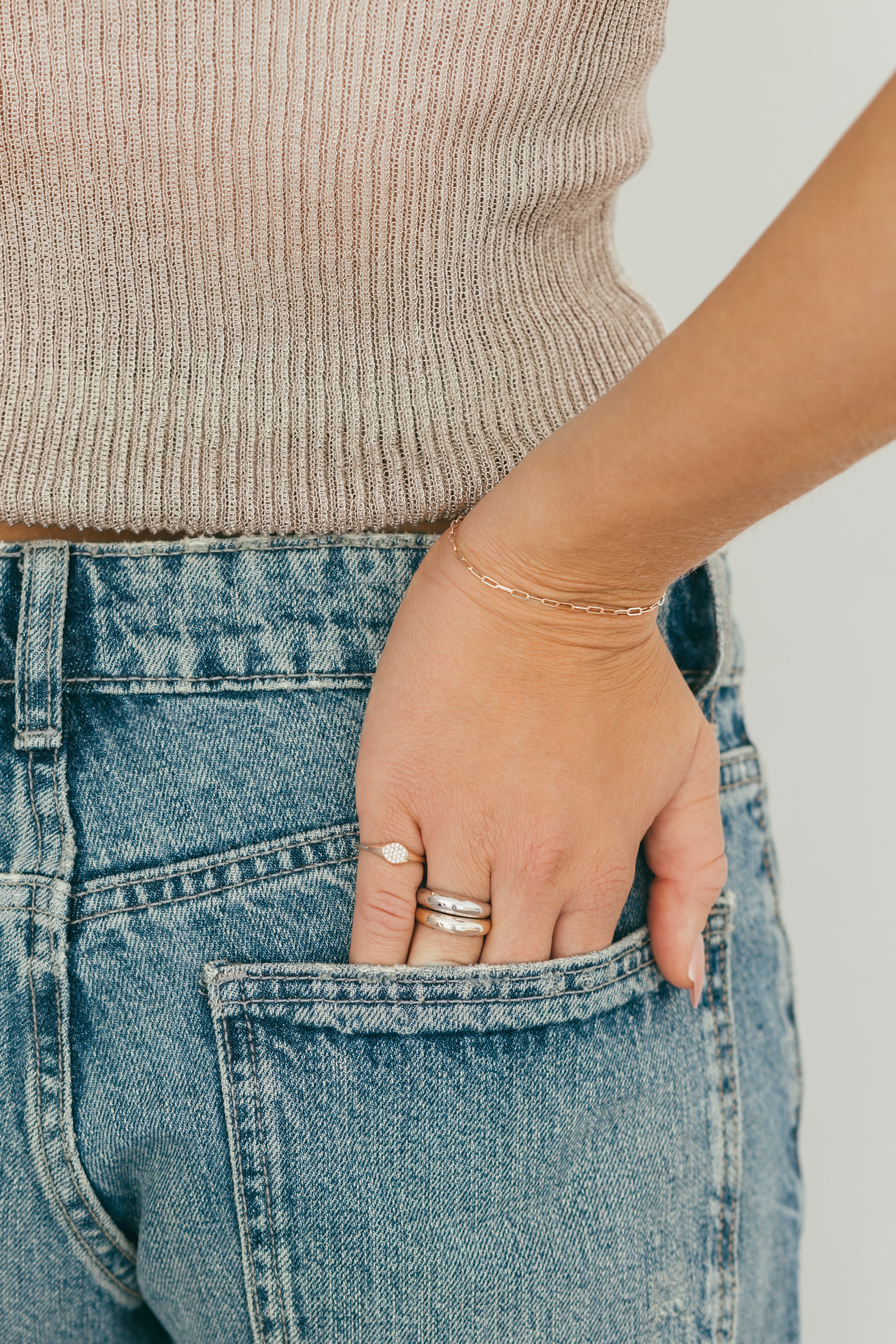 A person's hand accessorized with a mix of chunky gold and diamond rings, styled with a beige top and blue jeans on a neutral background.
