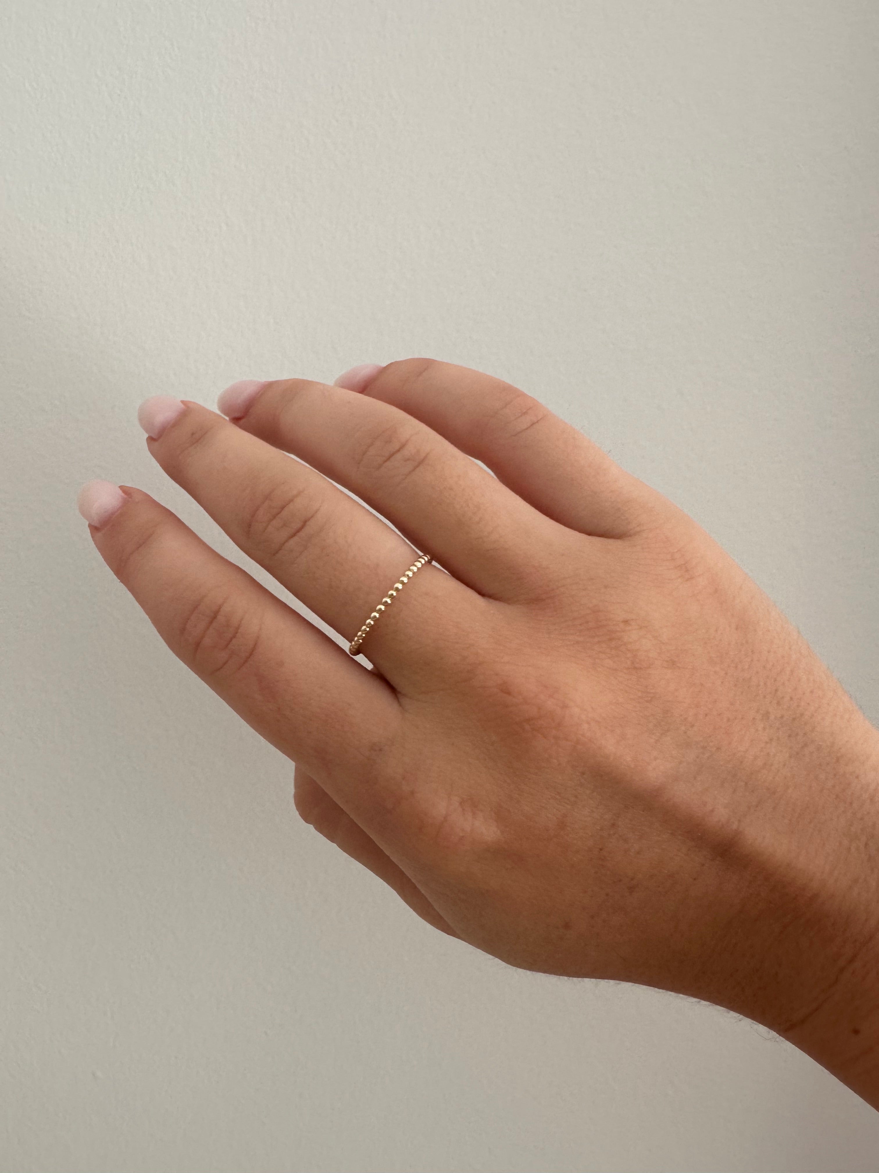 A gold delicate bead ring on a person's finger against a neutral background.