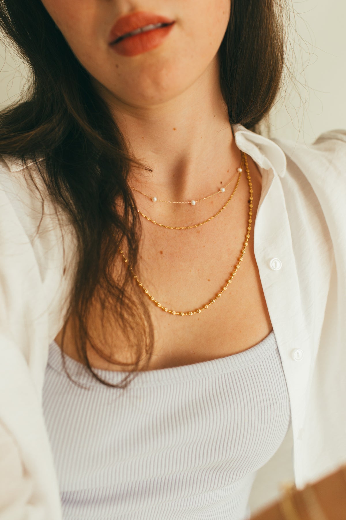 Close-up of a person wearing a stack of gold necklaces including a pearl and chain necklace, a twisted gold necklace and a bead gold necklace. 