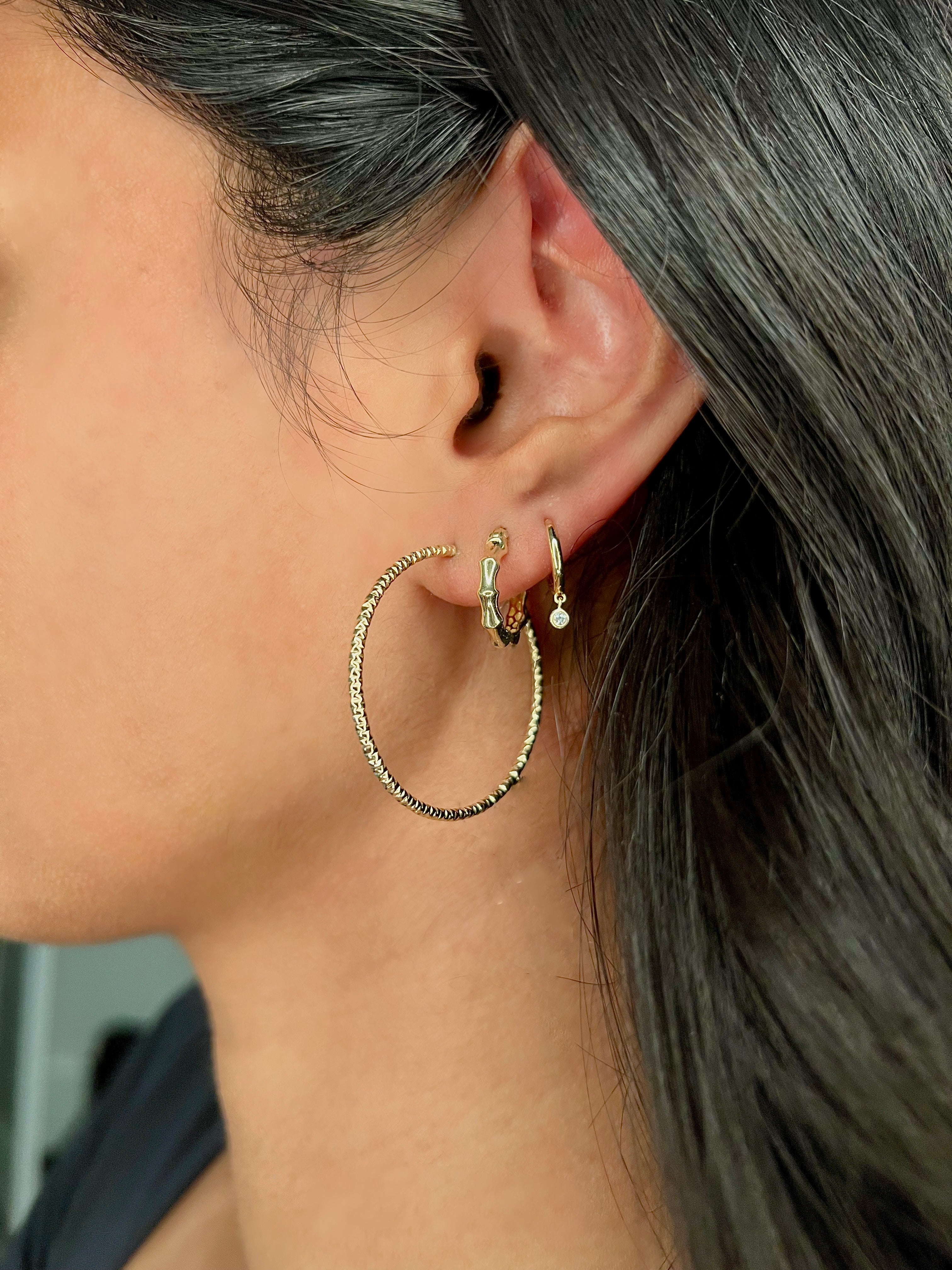 A close-up side profile of a woman wearing a trio of a gold diamond-cut hoop earring, a textured gold bamboo hoop, and a gold diamond bezel drop mini huggie earring.