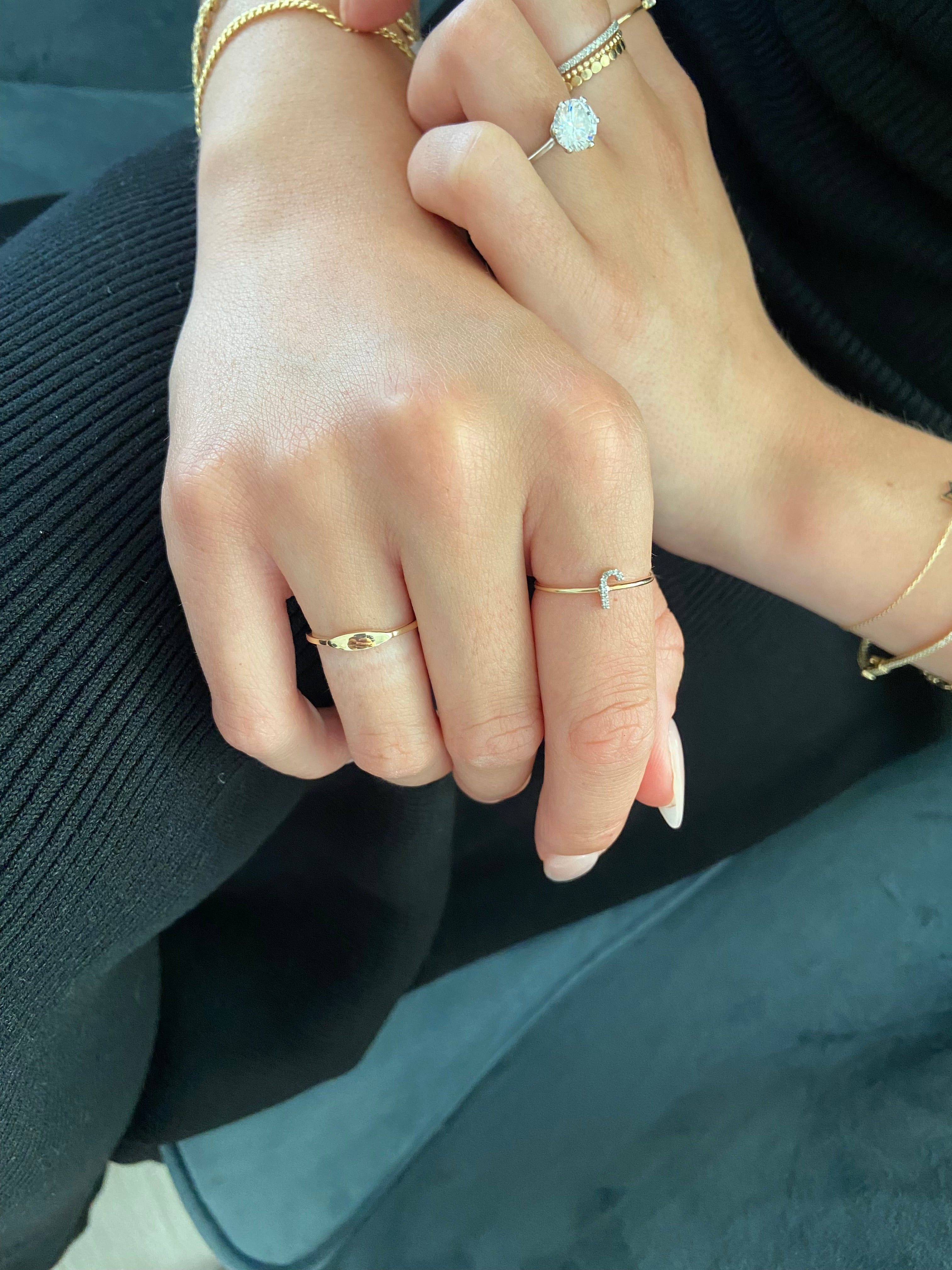 A person's hands styled with multiple gold rings including a mini oval signet ring and letter "J" ring against a dark background.