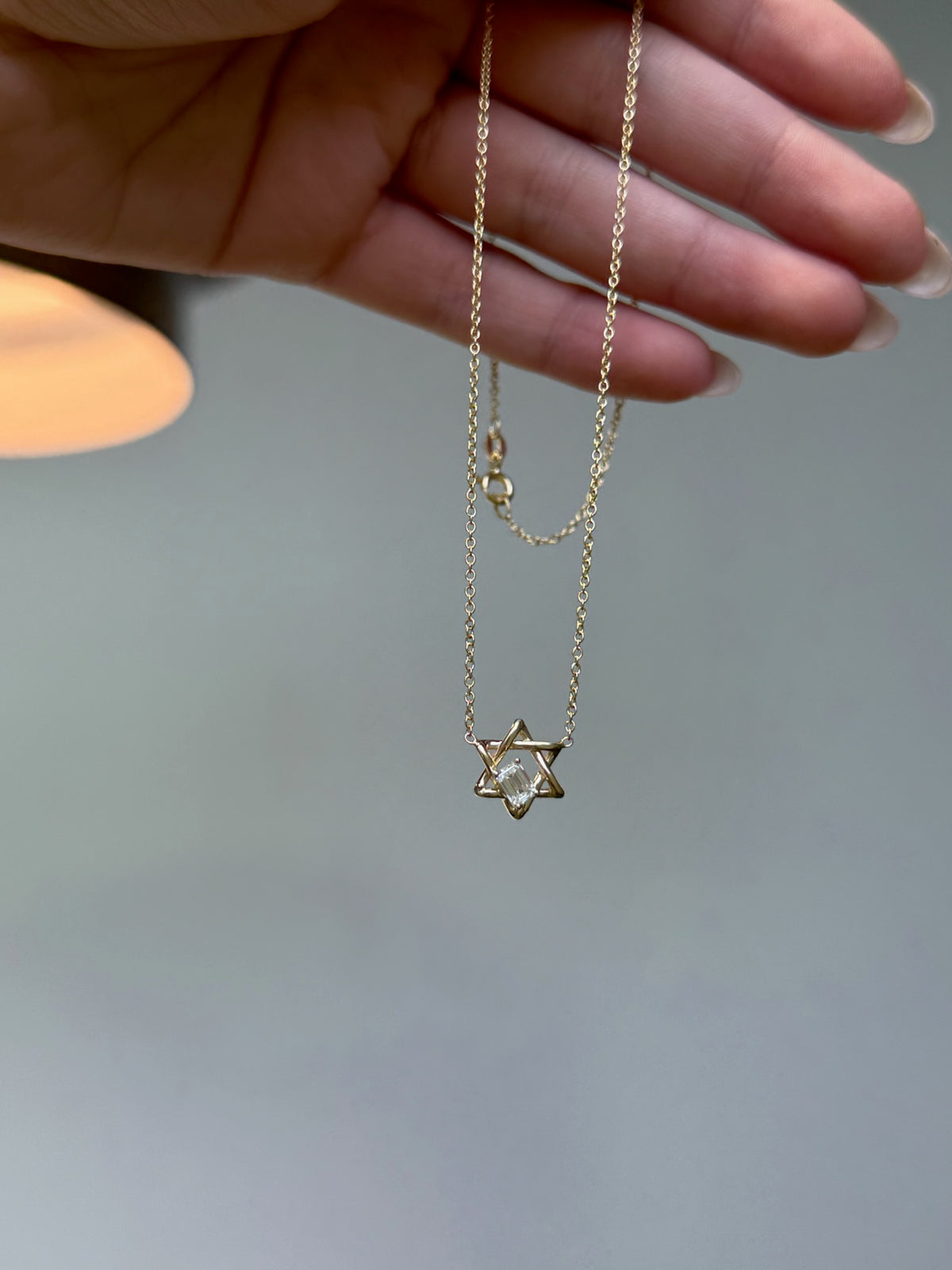 Gold necklace with a Star of David pendant held up by a hand, and featuring a floating emerald cut diamond in the center. 