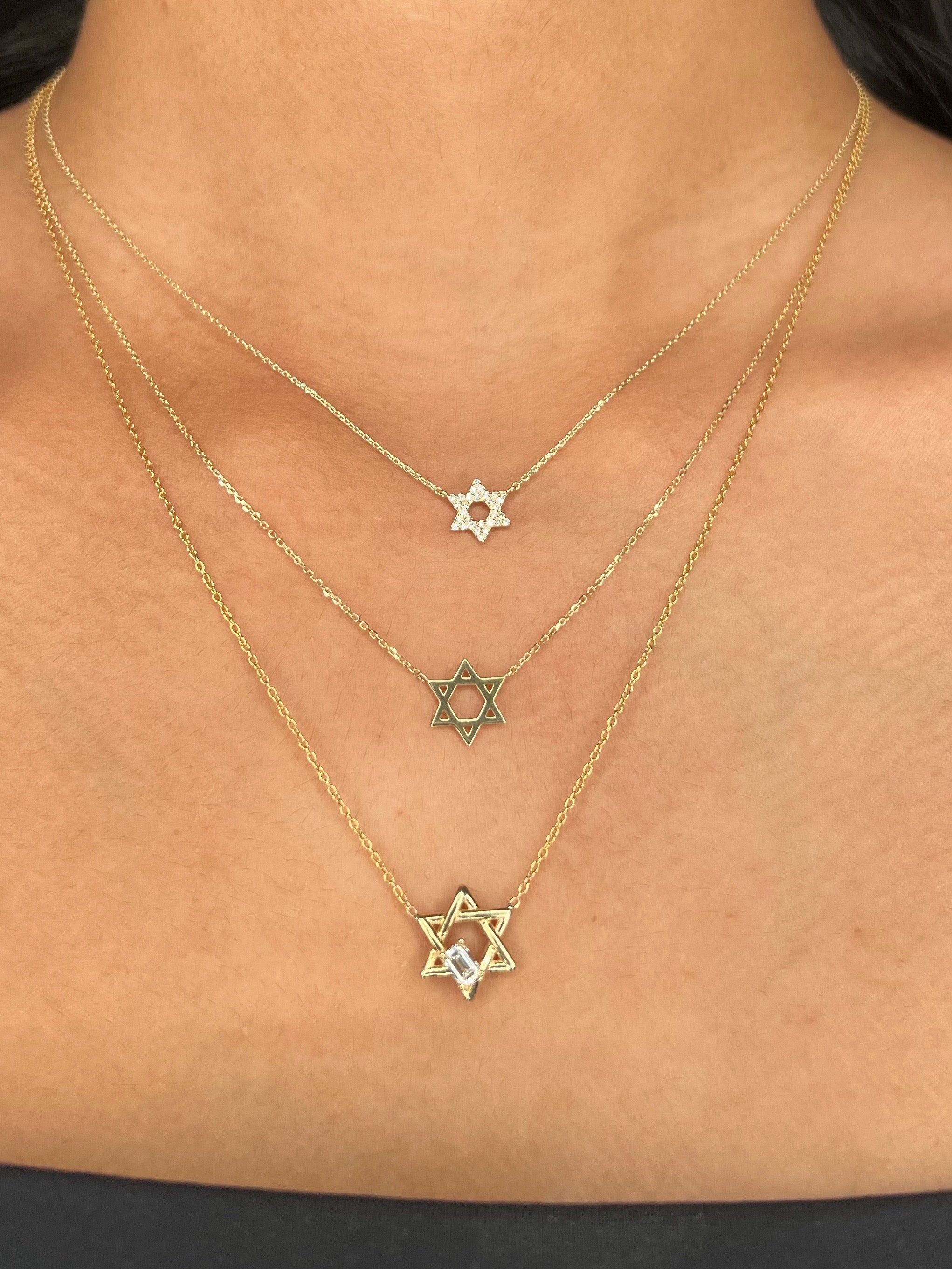 Three layered gold necklaces with Star of David pendants worn on a model's neck. The bottom necklace is an overlapping Star of David with a floating emerald diamond.