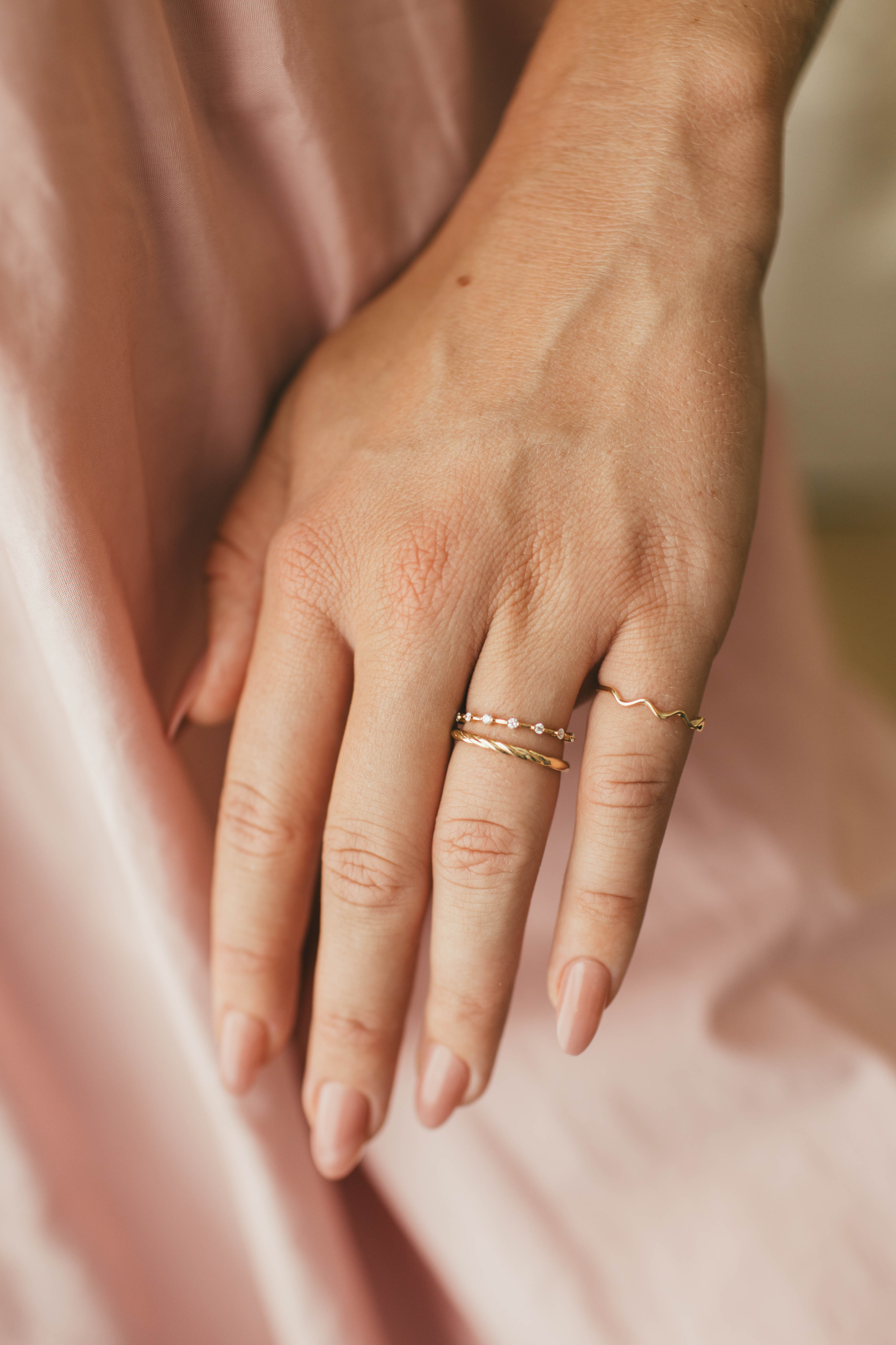 Hand wearing three gold rings on a soft pink fabric background.