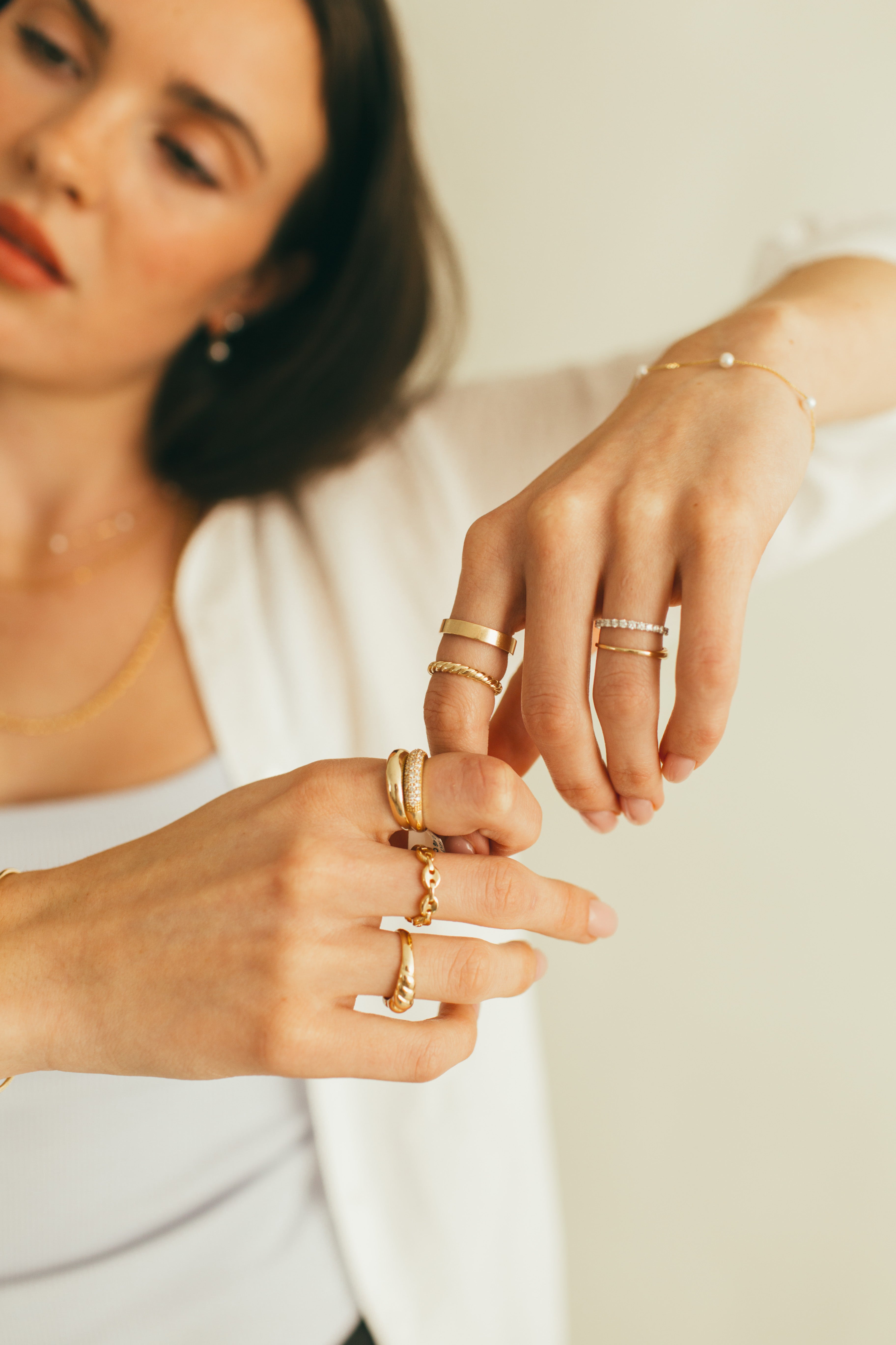 A woman's hands styled with a mix of chunky, thin, and flat gold bans and diamond rings on a blurred background.