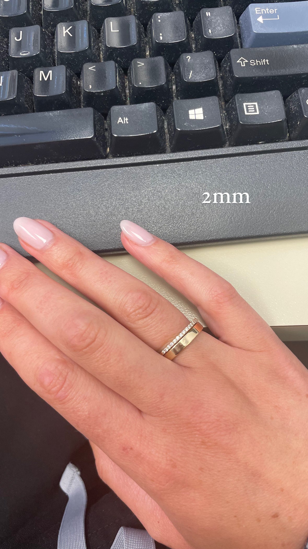 A person's hand resting on a black keyboard, wearing a thin diamond ring stacked on top of a gold wide flat cigar band, with text indicating a "2mm" ring width.