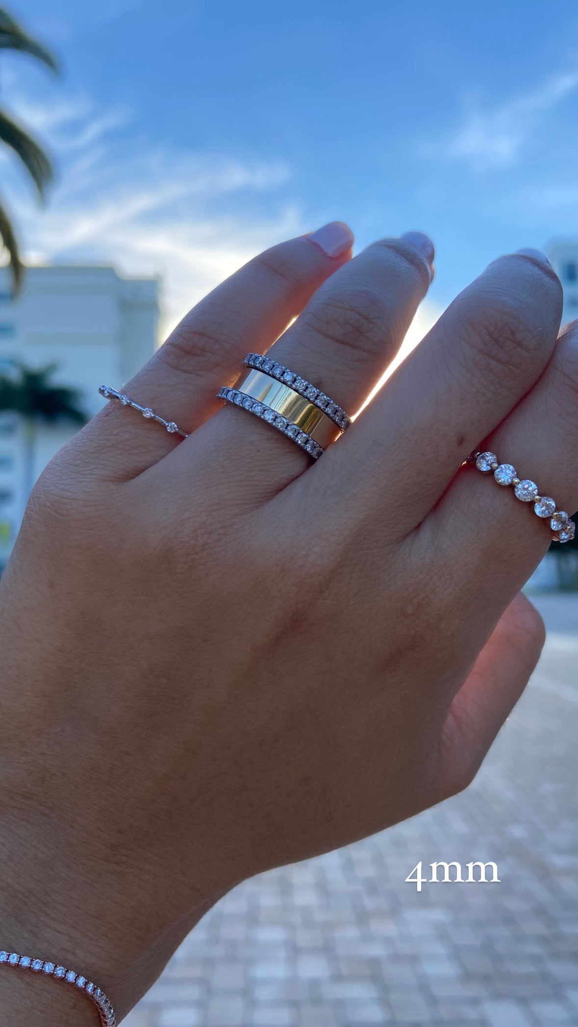 A close up of a person's hand wearing multiple diamond rings and a gold wide flat cigar band on a backgorund with a blue sky and palm trees, with text indicating a ring width of "4mm".