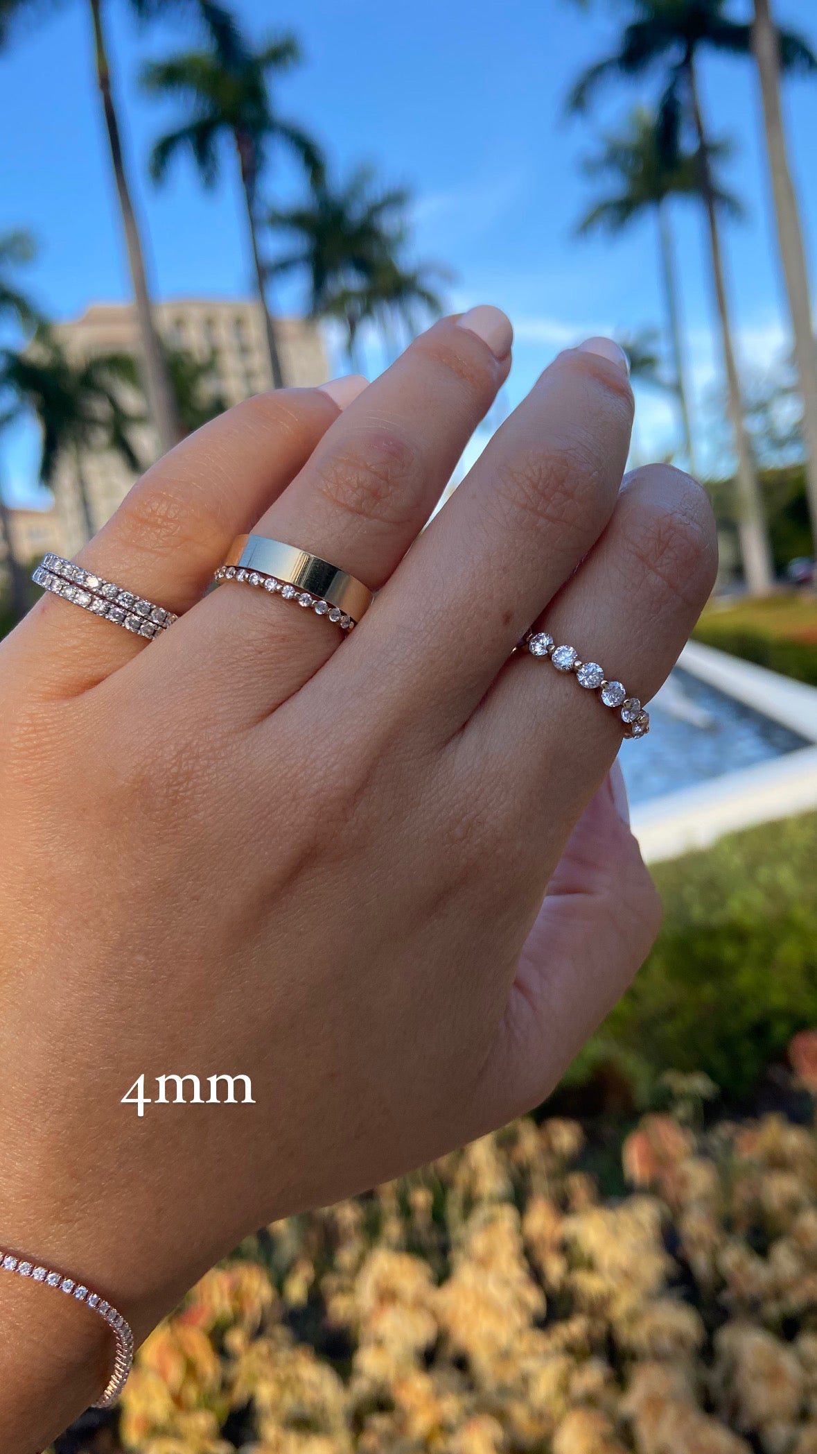 A close up of a person's hand wearing multiple diamond rings and a gold wide flat cigar band, on a background with a blue sky and palm trees, with text indicating a "4mm" ring width.