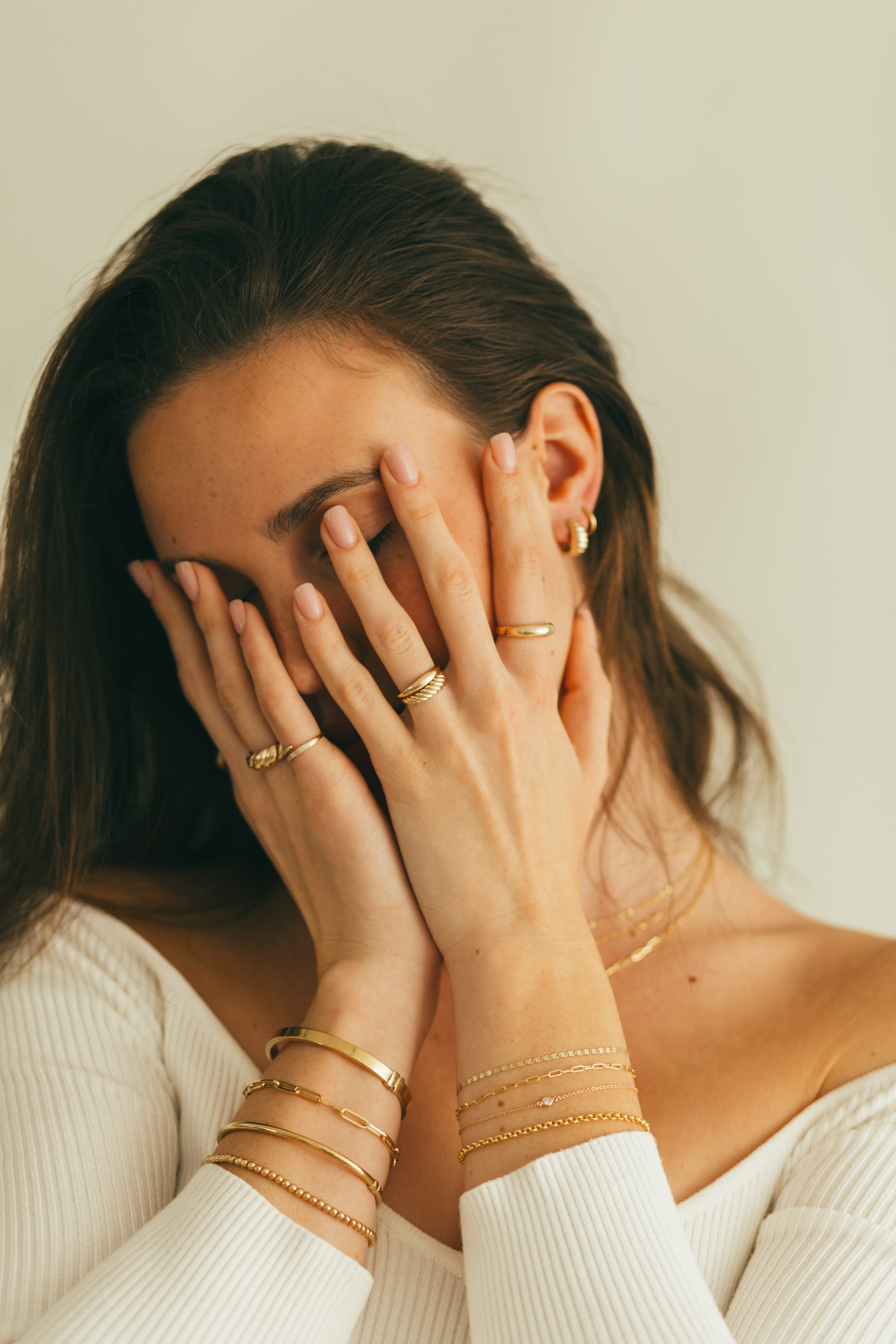 A woman wearing gold jewellery, including a variety of elegant rings and multiple bracelets covering her face against a neutral background.