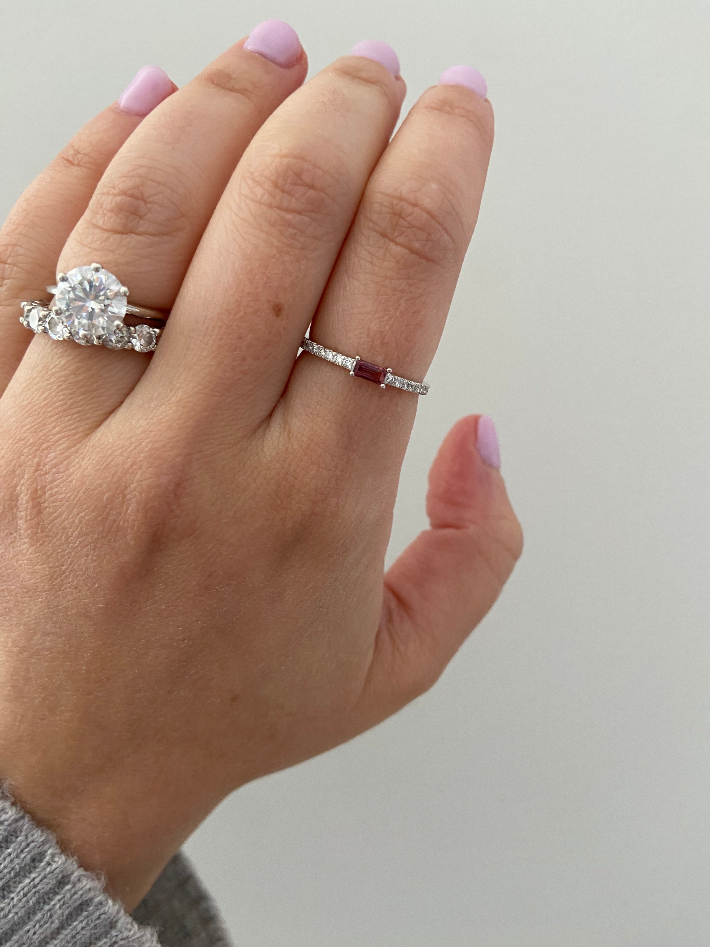 Close-up of a hand wearing a baguette birthstone and diamond ring with other diamond rings on a neutral background.