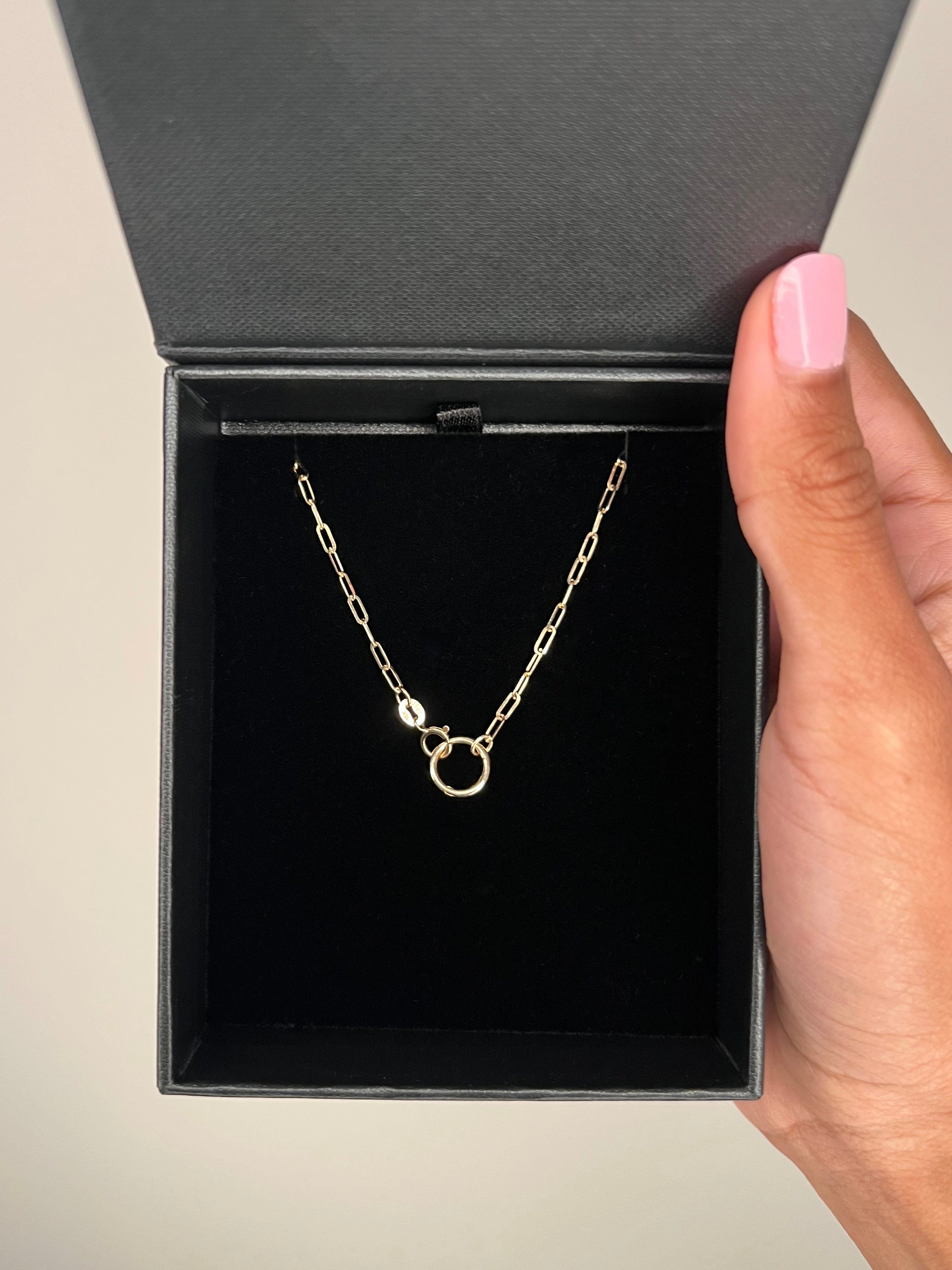 A 14K Gold Charm Connector attached to a gold paperclip chain in a black cushioned box held against a neutral background.