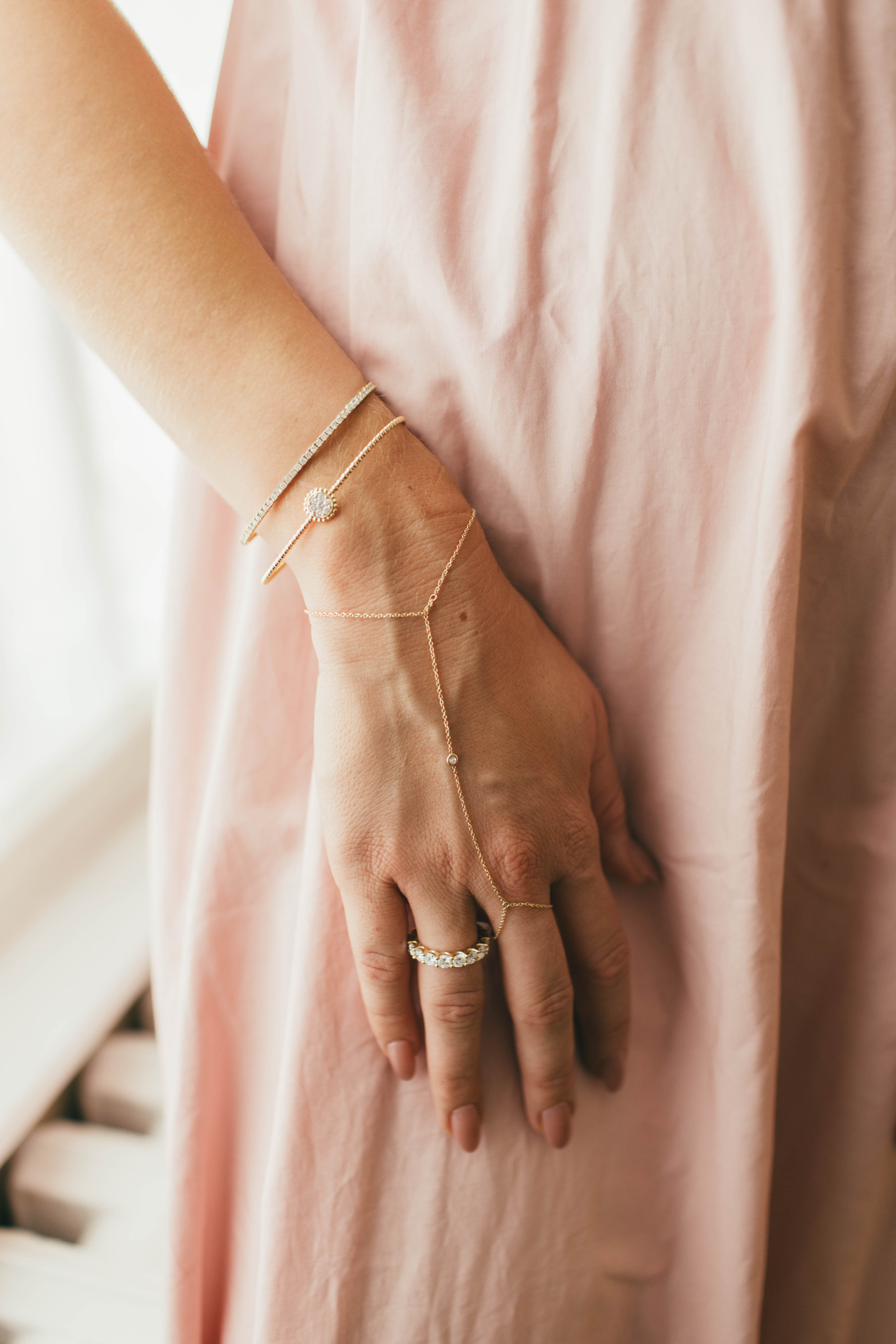A hand with a gold diamond hand chain, gold bracelets, and a ring on a soft pink fabric background.