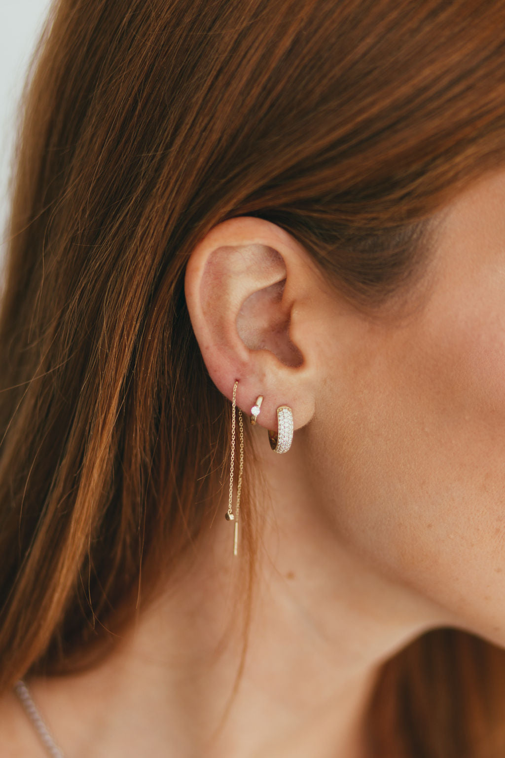 A close-up side profile of a woman wearing delicate gold and diamond hoop earrings with a thin threader chain against a clean white background.