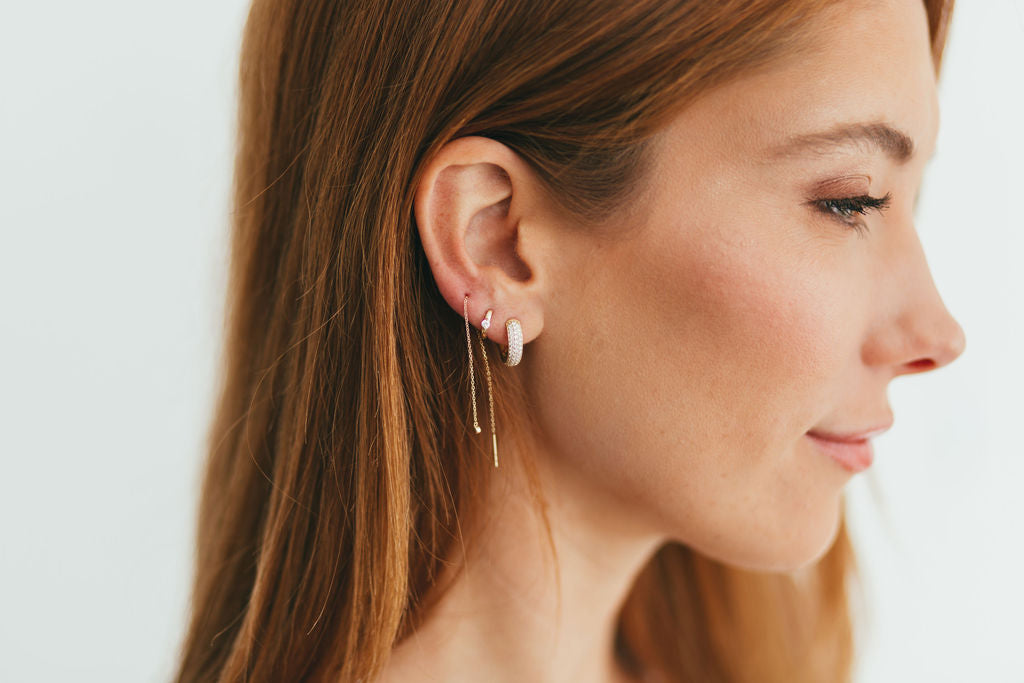 A close-up side profile of a woman wearing delicate gold and diamond hoop earrings with a thin threader chain against a clean white background.