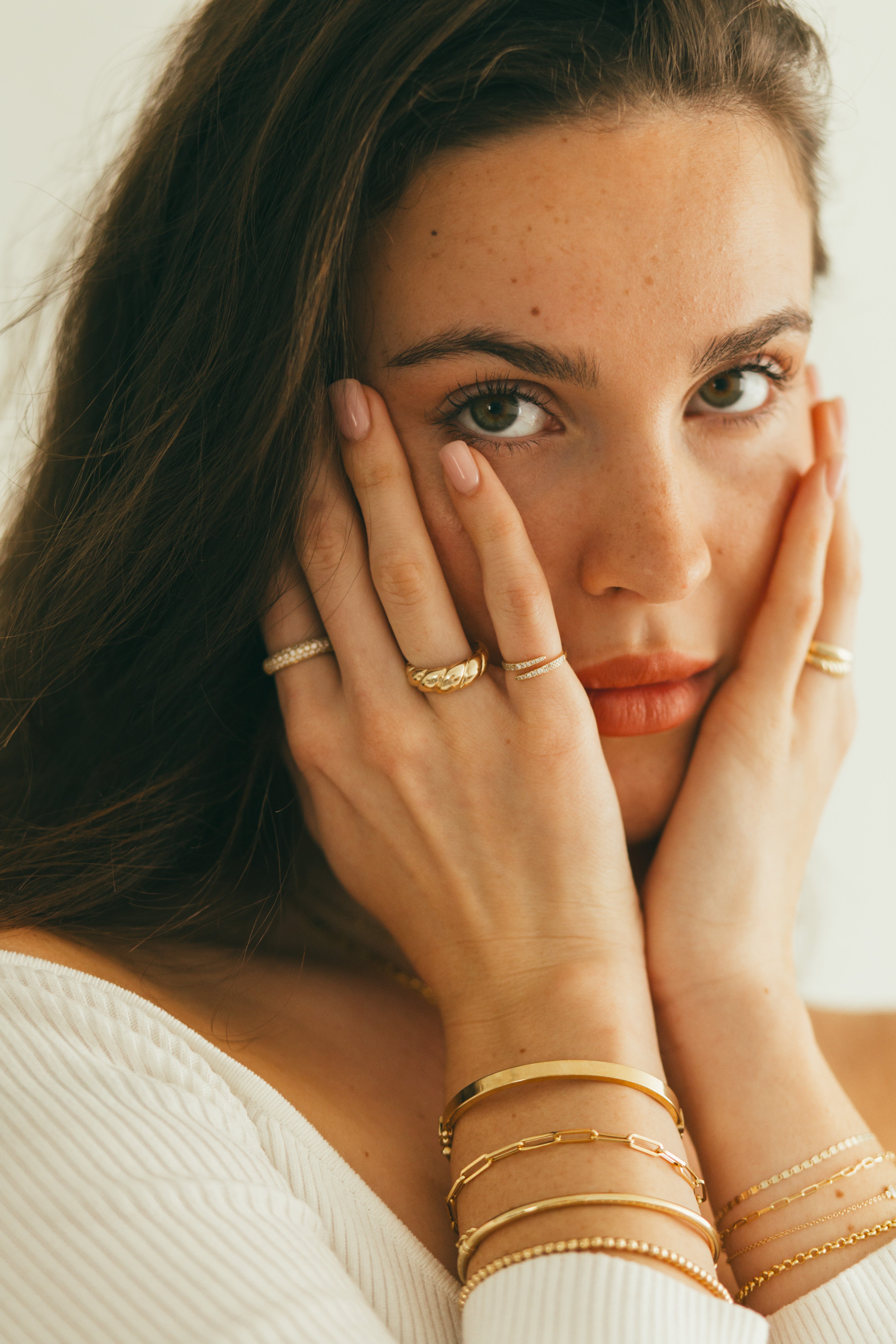 A woman holding her face with her hands, accessorized with a mix of thin and chunky gold rings and stacks of gold bracelets.