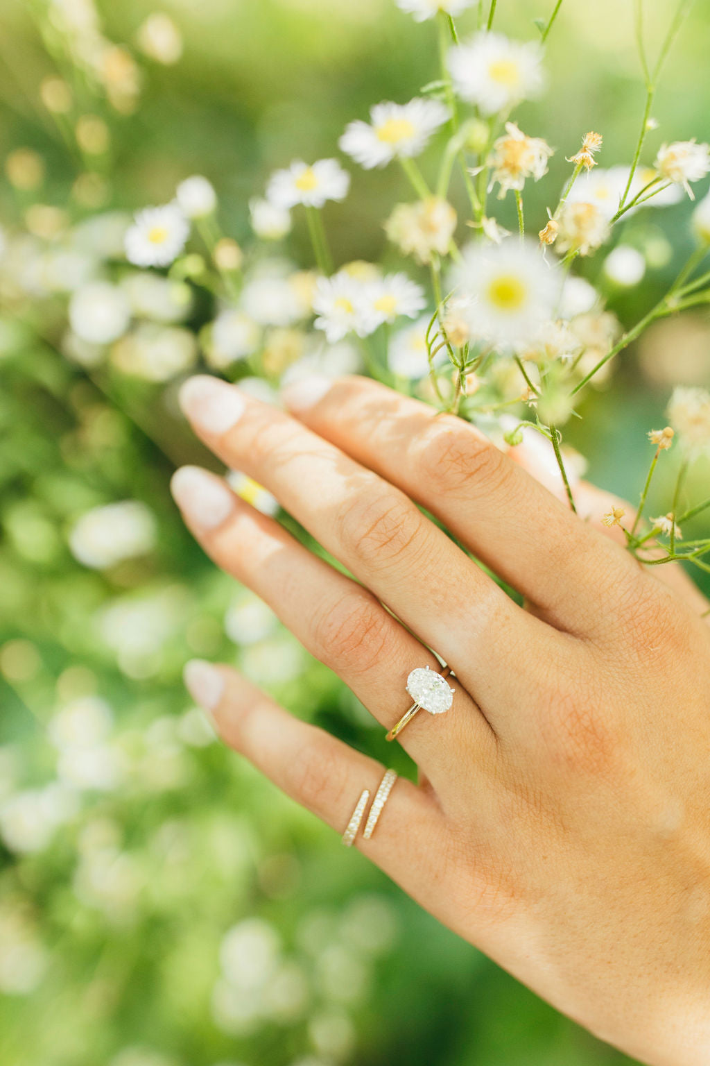 A hand styled with two gold and diamond rings on a blurred background of greenery and flowers.