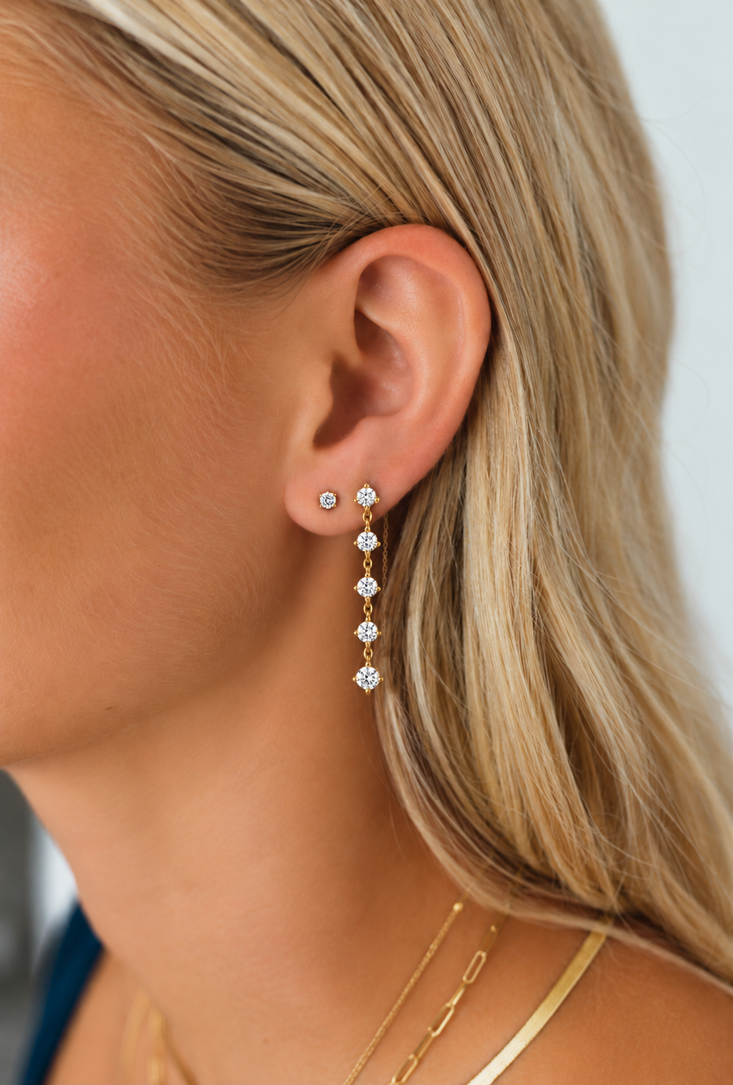 A close up of a model's side profile, wearing a diamond chain earring and simple diamond stud