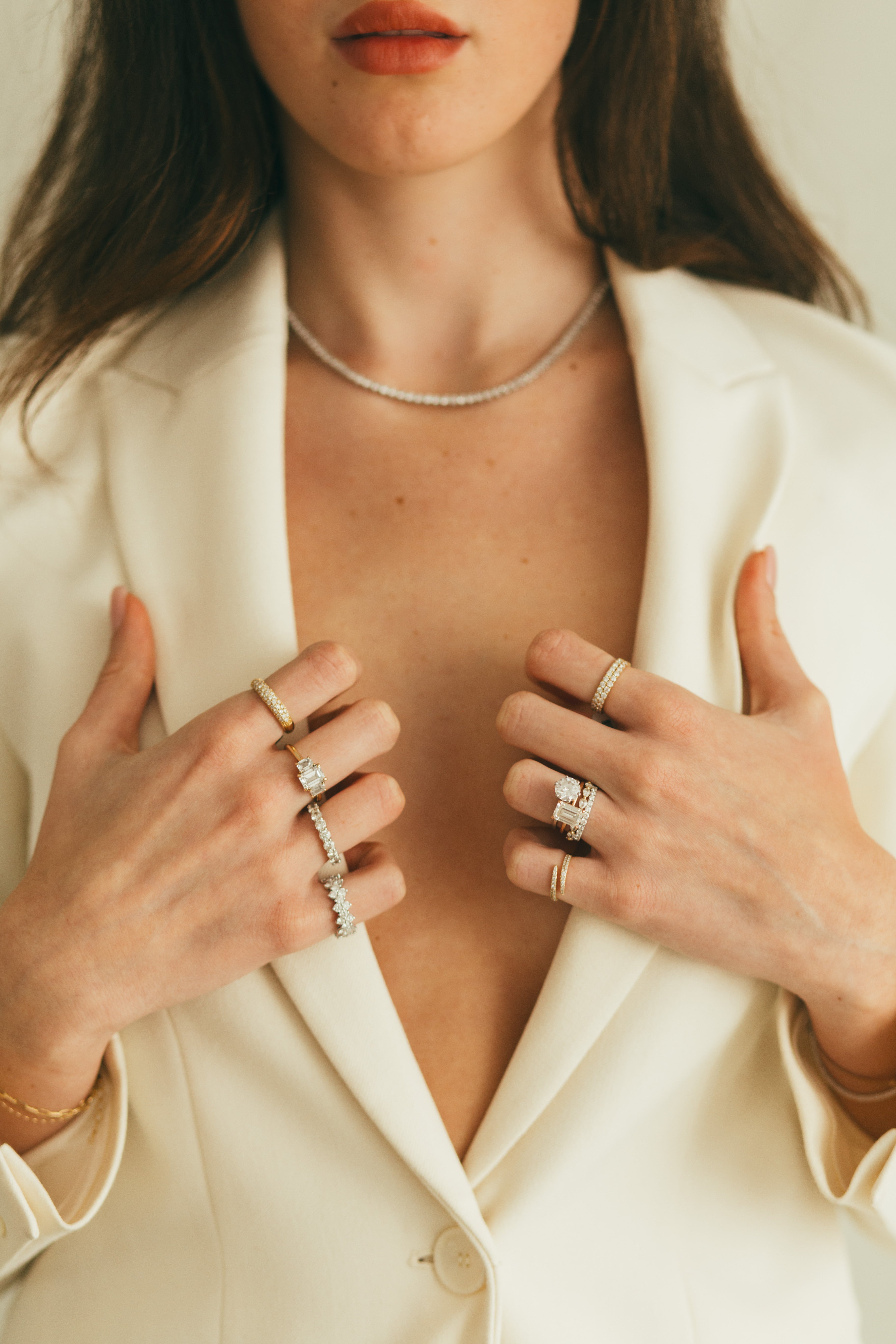 Close-up of a woman wearing multiple diamond rings, holding her off-white blazer, on a neutral background.