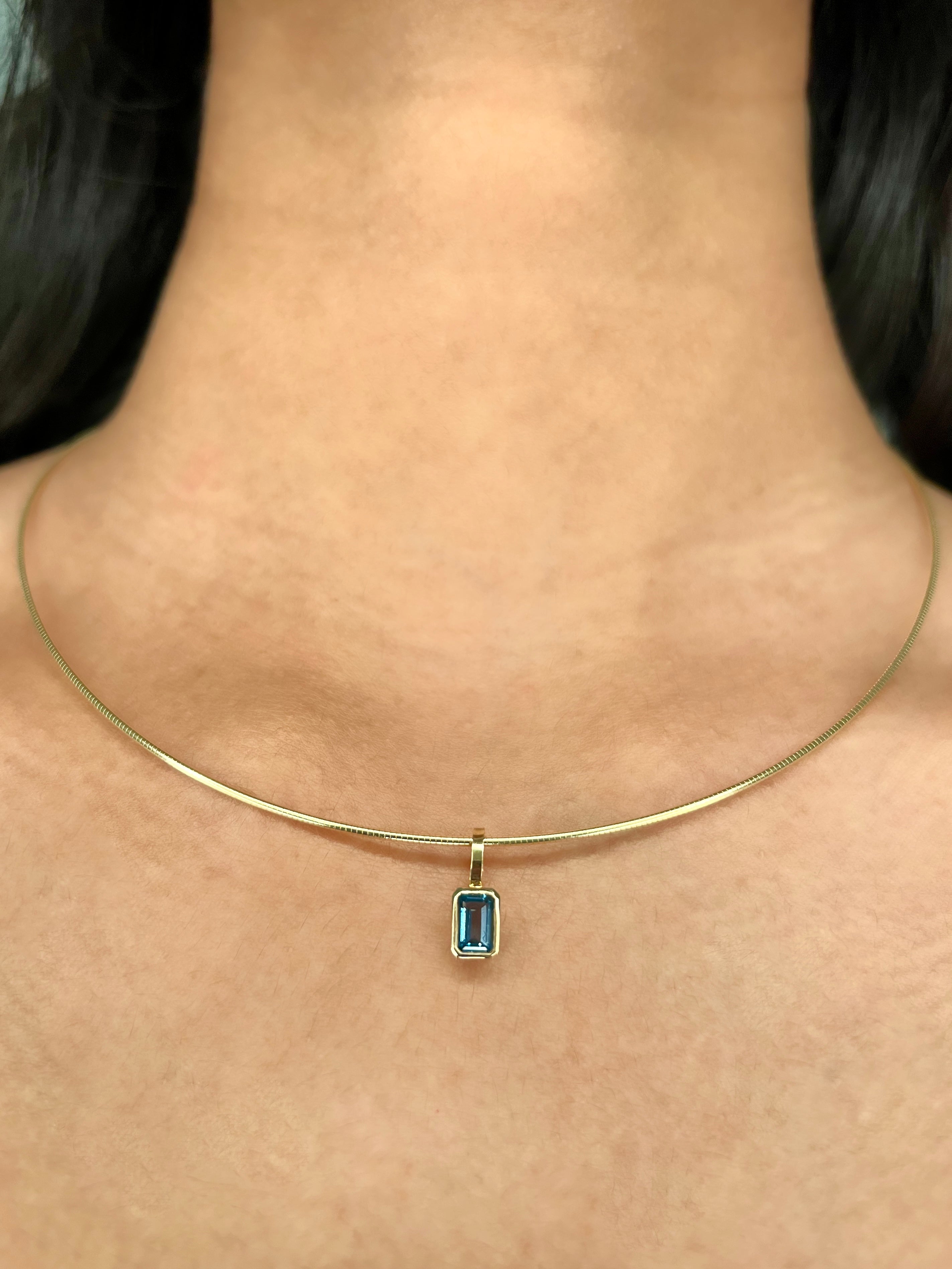 A close-up of a delicate gold solid diamond-cut wire necklace worn on a person's neck, featuring a small rectangular bezel-set emerald-cut birthstone charm.