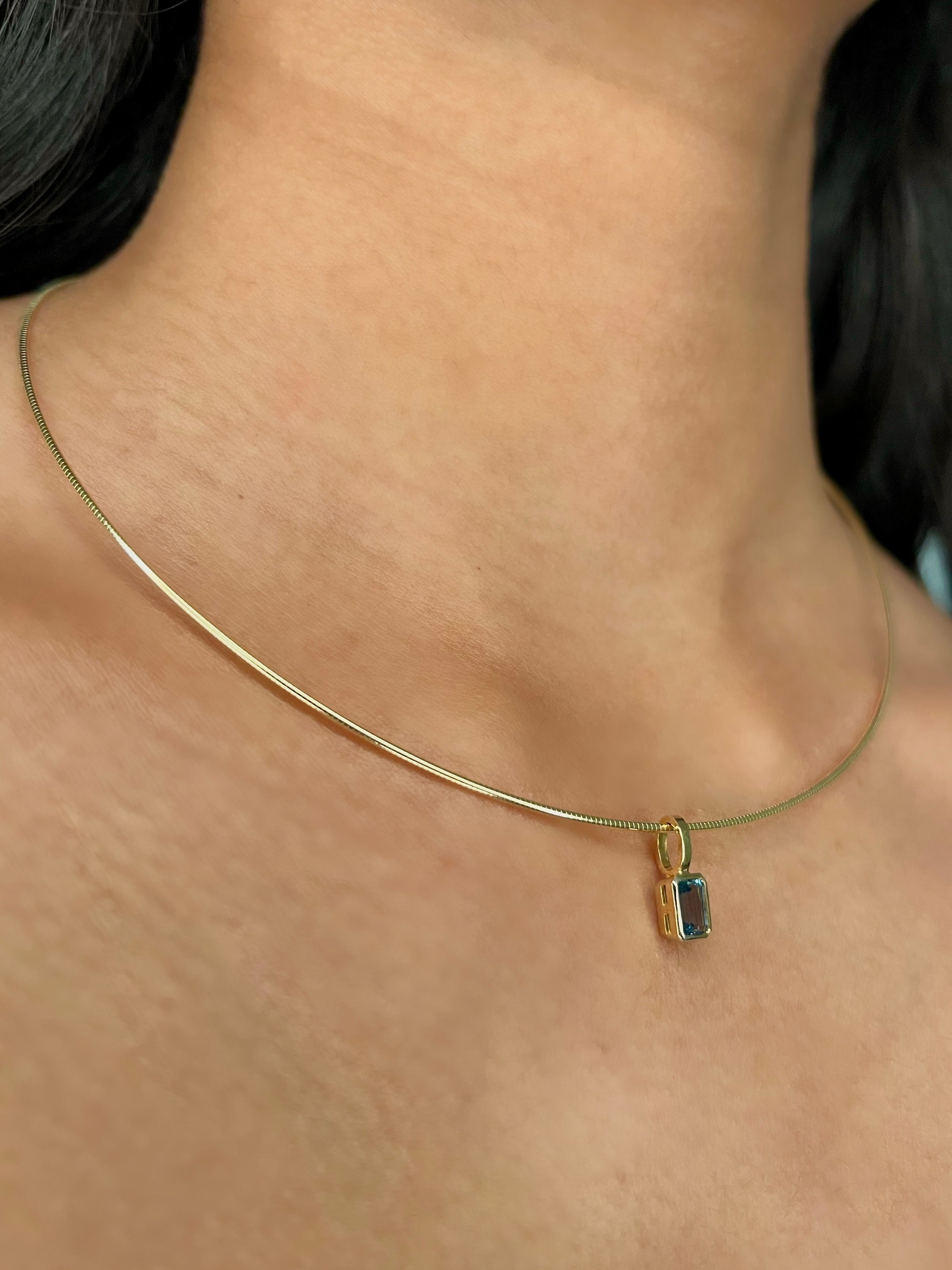 A close-up of a delicate gold solid diamond-cut wire necklace worn on a person's neck, featuring a small rectangular bezel-set emerald-cut birthstone charm.